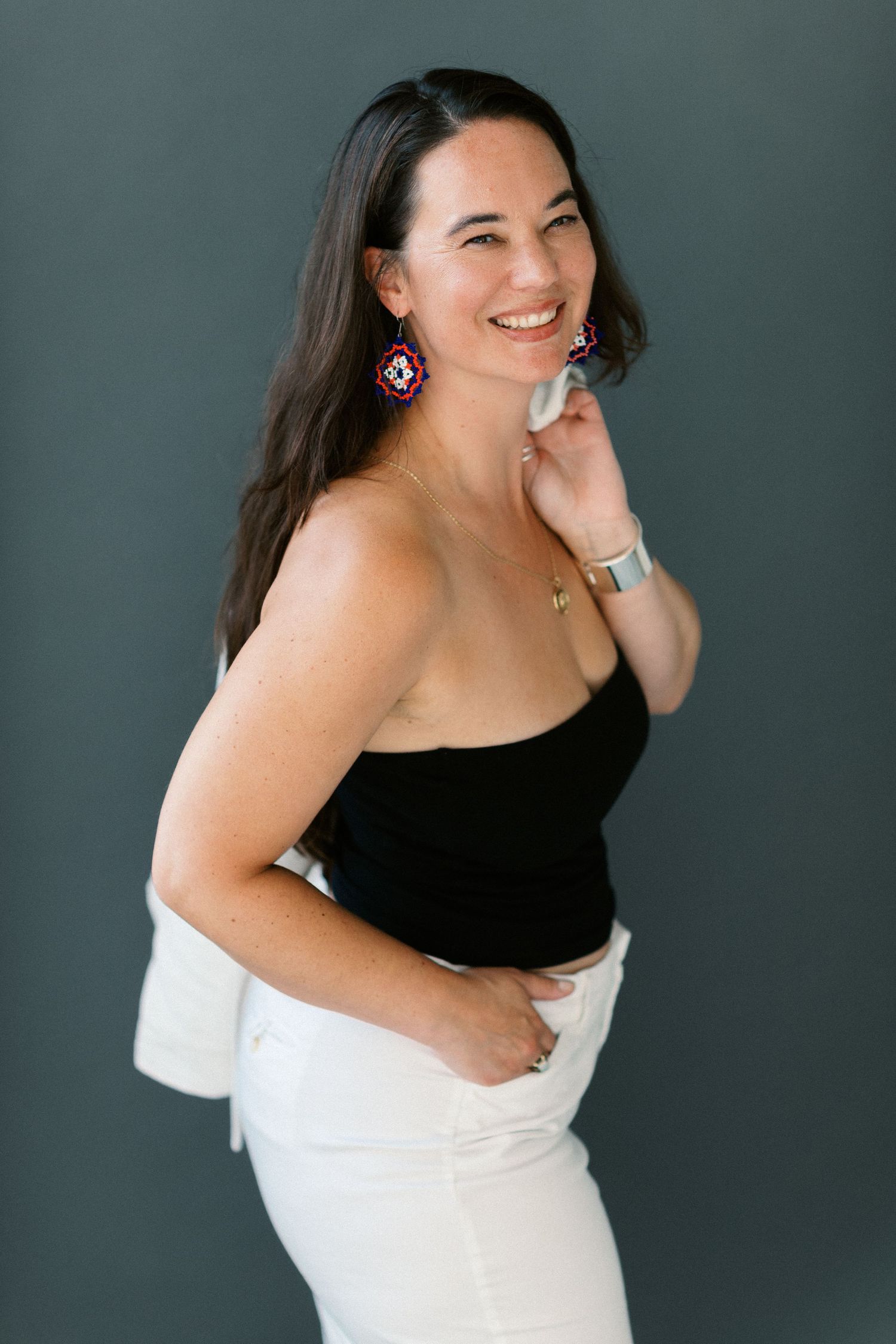 Smiling woman in black top, white pants, and colorful earrings posing against a gray background.