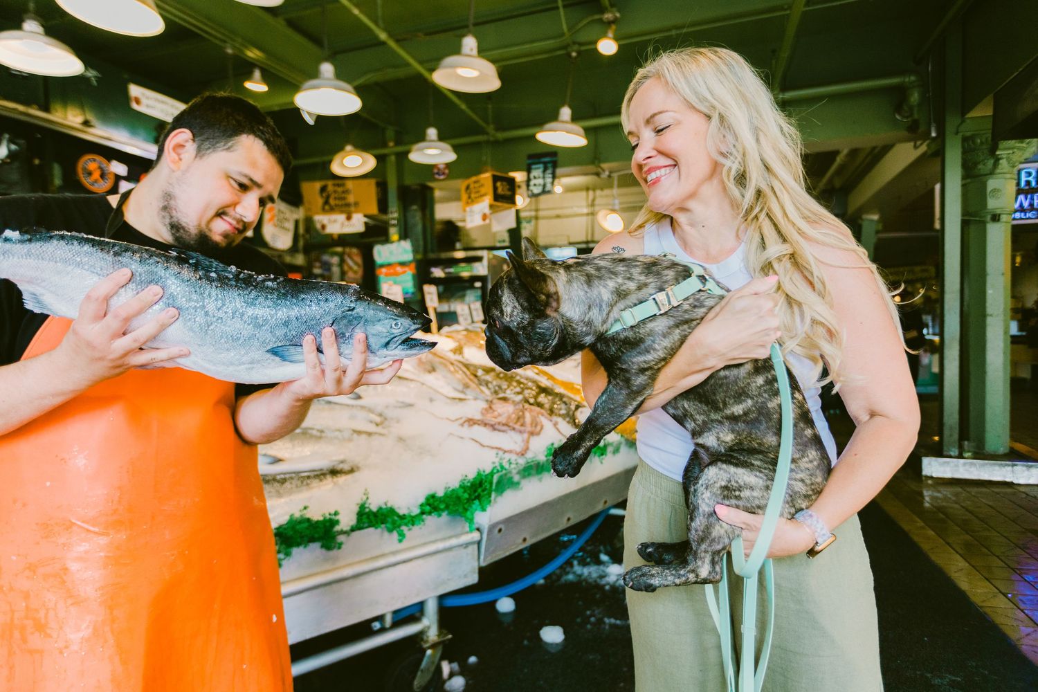 Man and woman laughing while holding large fresh fish at a seafood market counter.