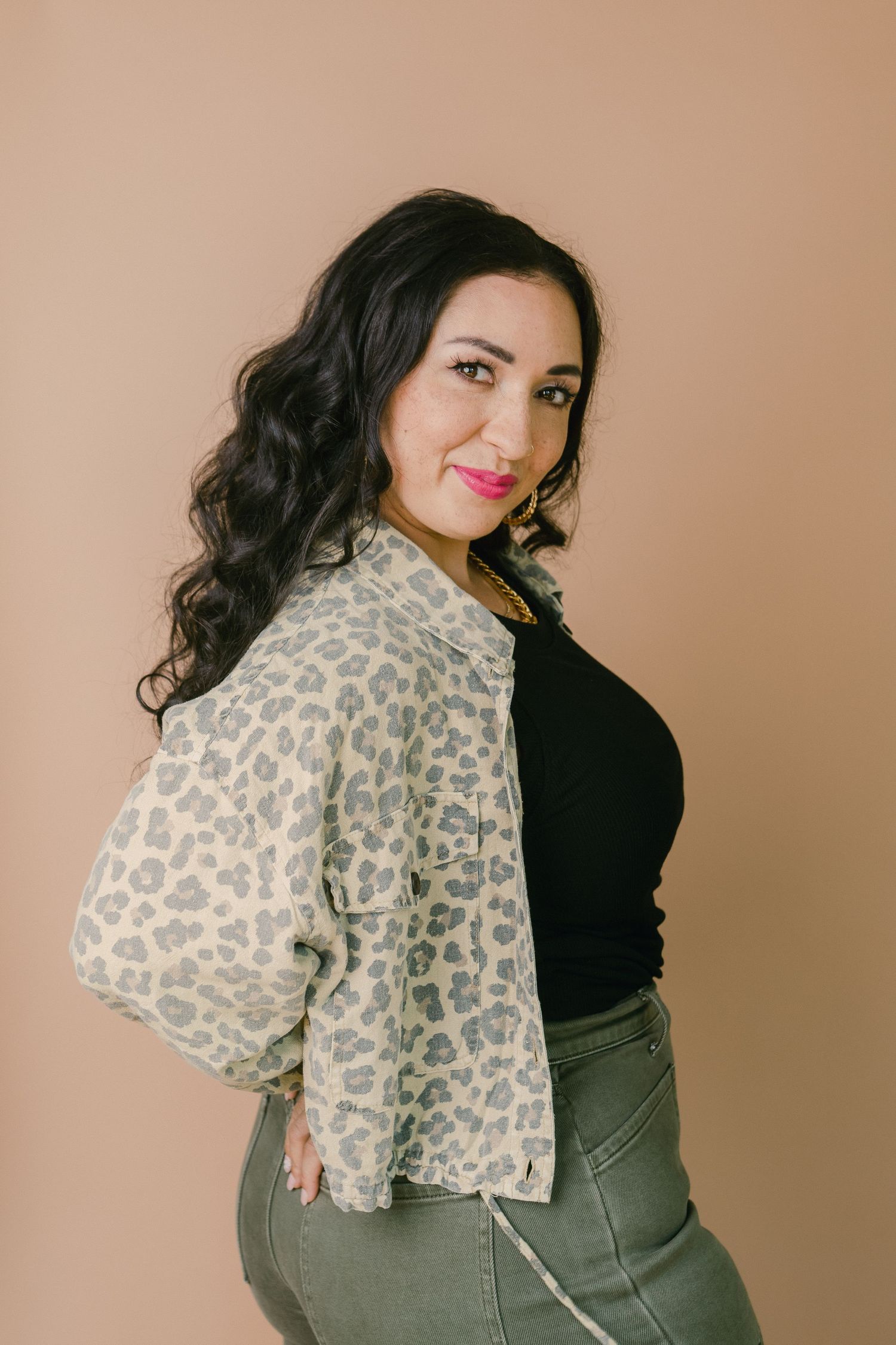 Woman in leopard print jacket, black top, and olive jeans poses against a beige background with curly dark hair.