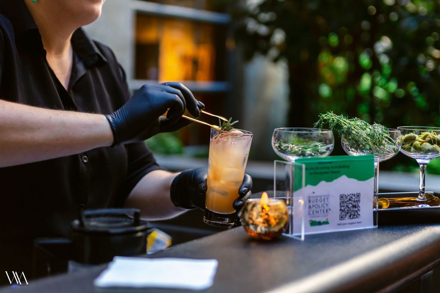 Bartender in black gloves garnishing a craft cocktail with herbs at an outdoor bar.