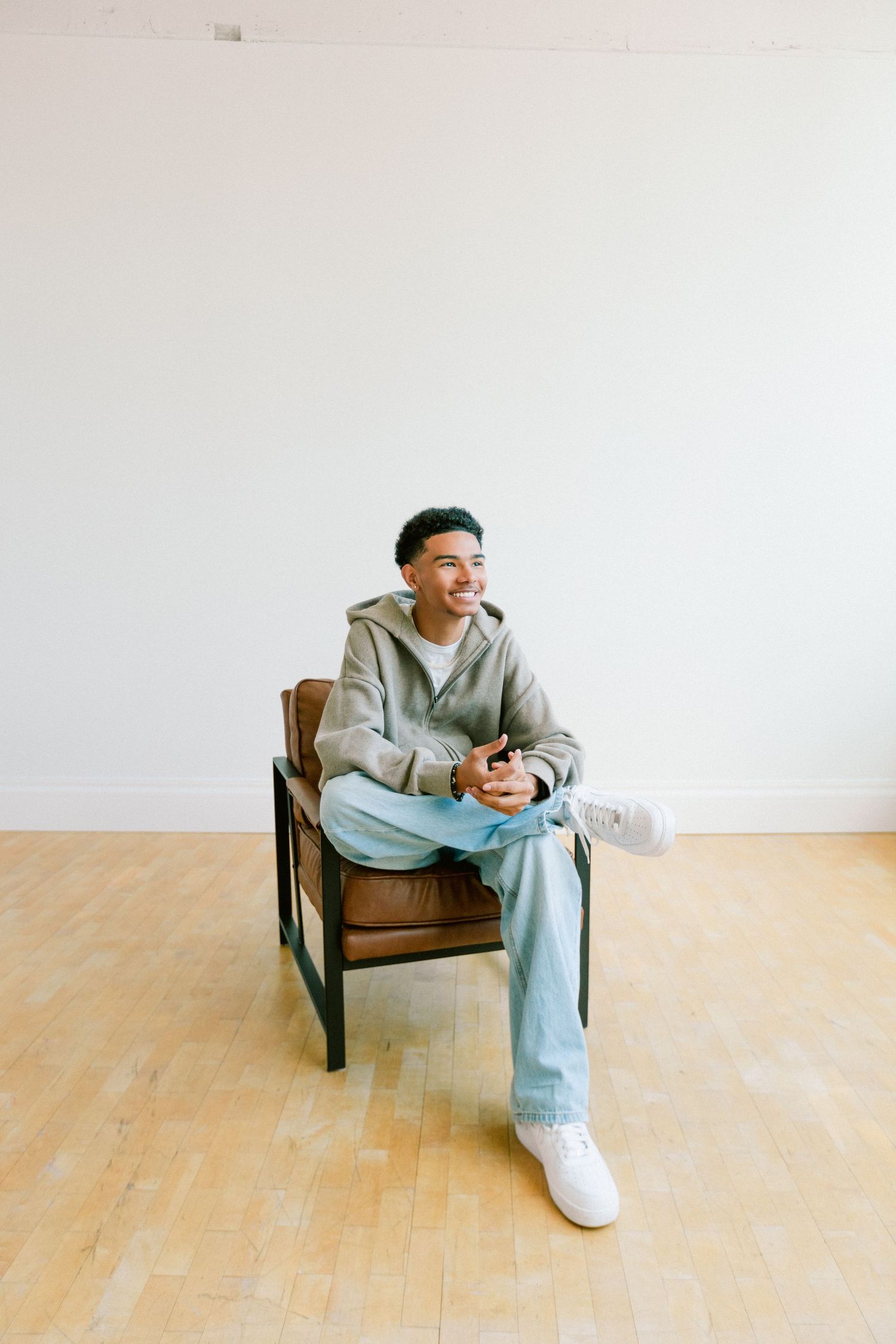 Young man in gray hoodie and jeans sitting relaxed in brown leather chair on hardwood floor, smiling and holding phone.
