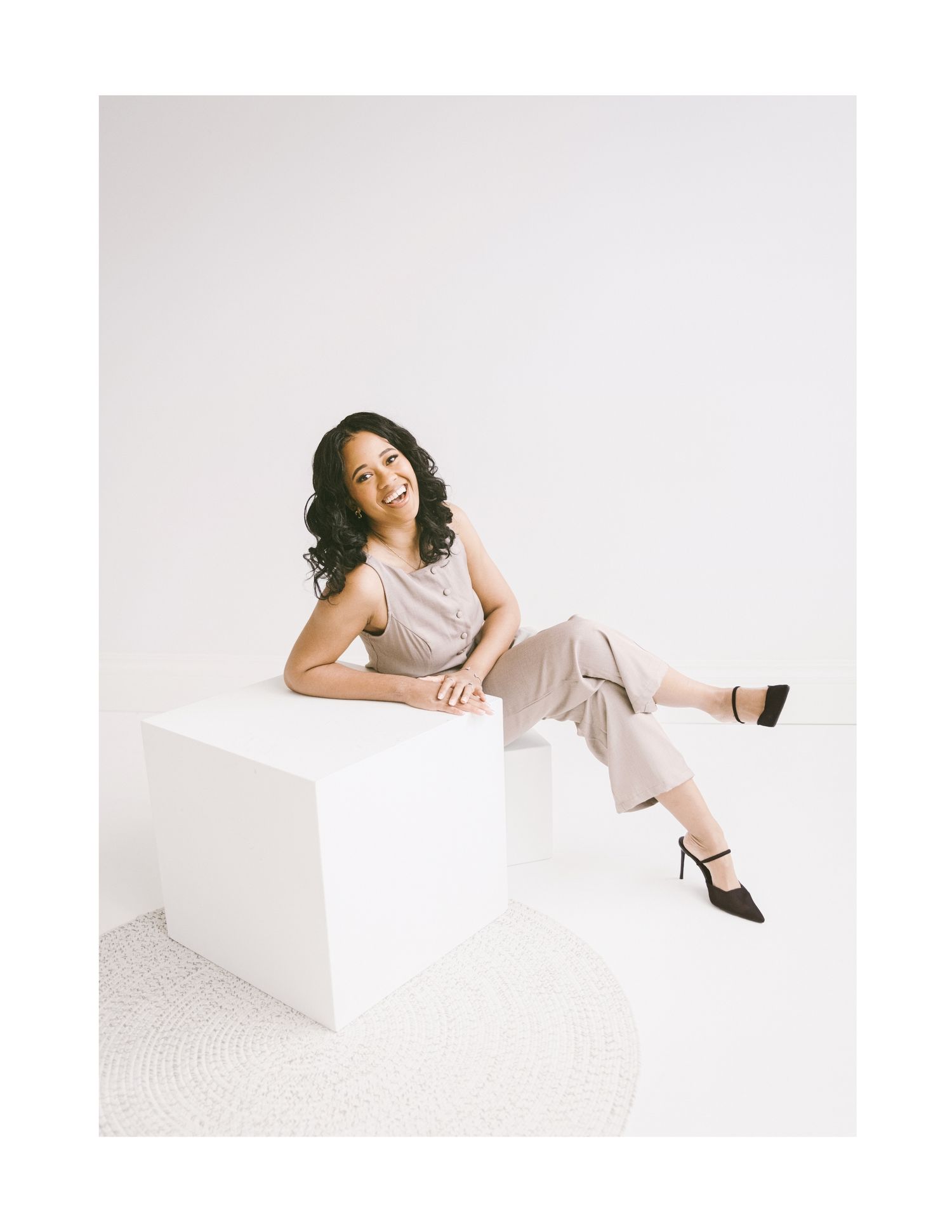 A smiling woman in a light pink jumpsuit and black heels sits gracefully on a white cube against a white background.