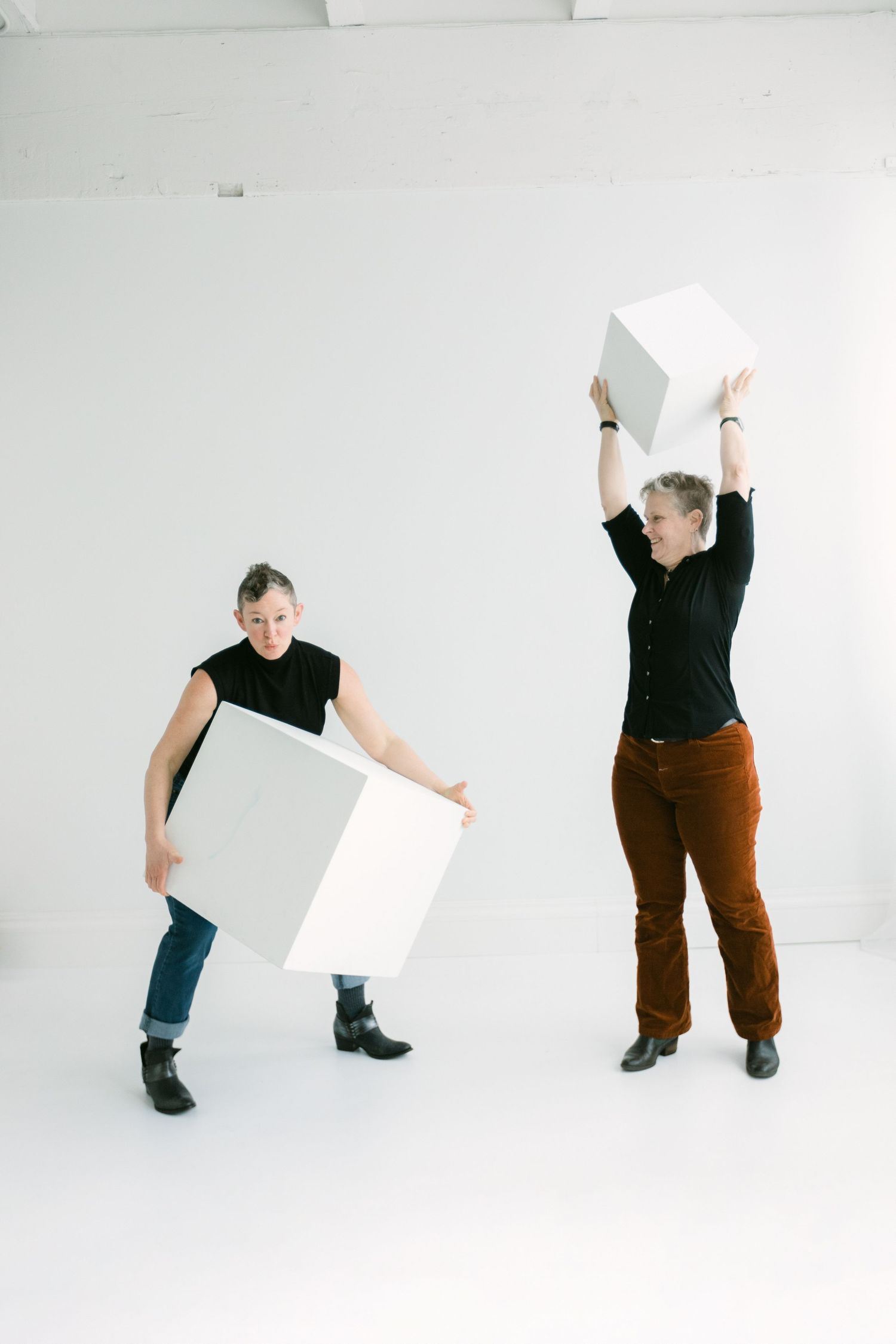 Two women in black tops playfully holding white geometric cube boxes in a bright white studio space.