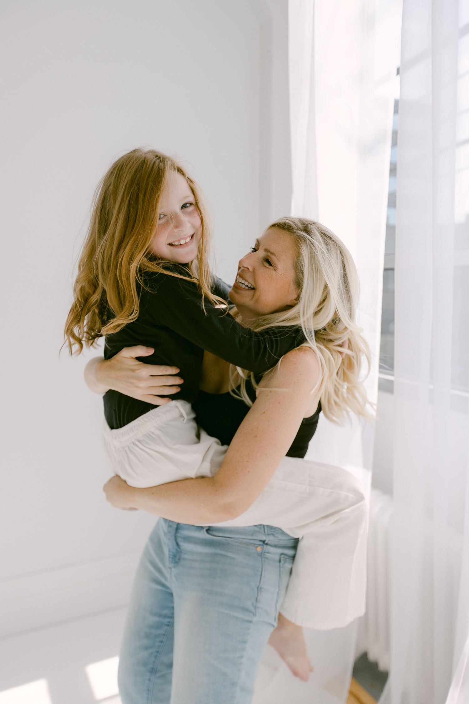 A blonde woman joyfully lifts a smiling redheaded girl in a bright white room near a window.