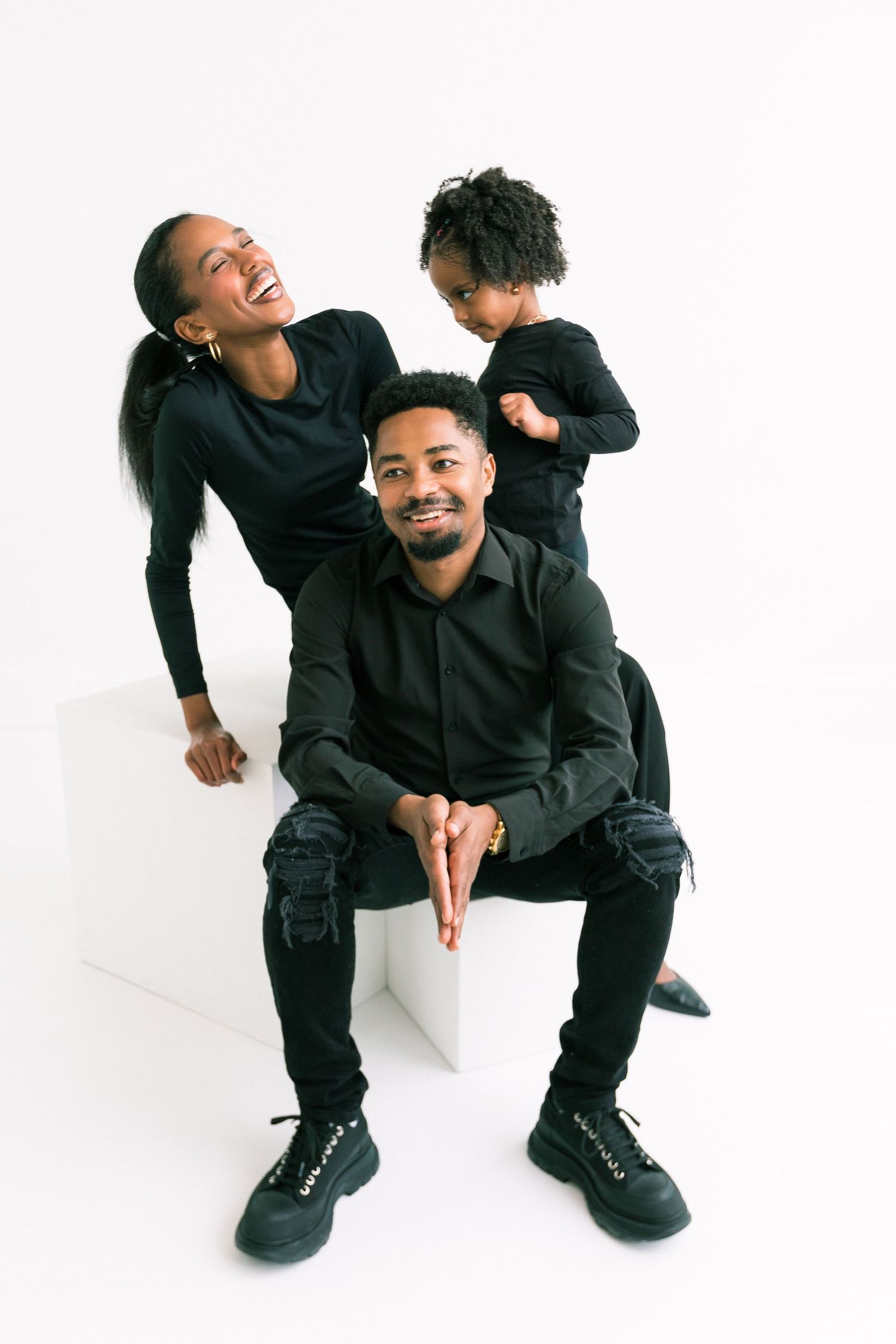 A joyful Black family of three in matching black outfits poses together against a white background in a studio photo.