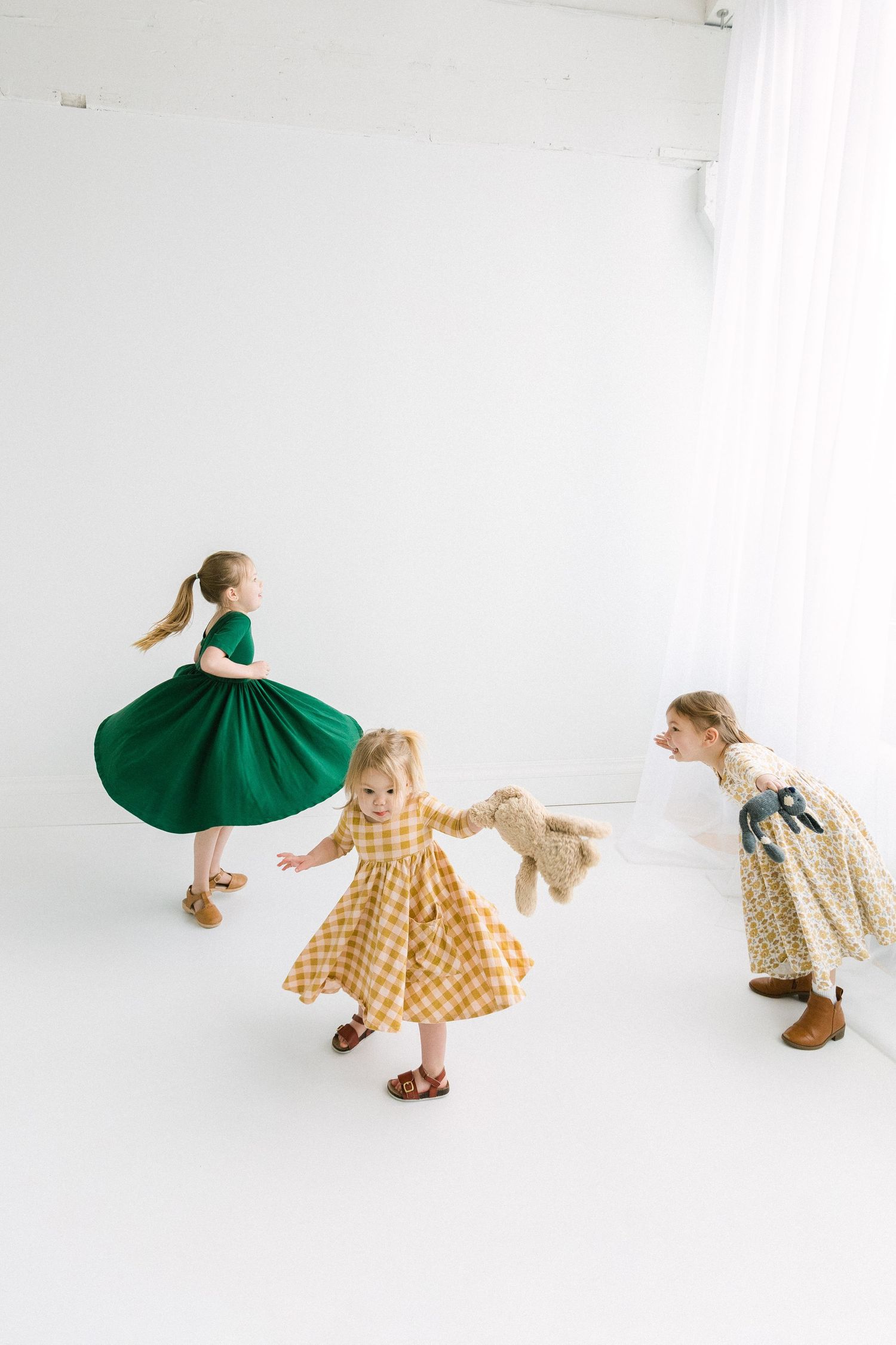 Three young girls in colorful dresses play together in a bright white studio, one twirling in a green skirt.
