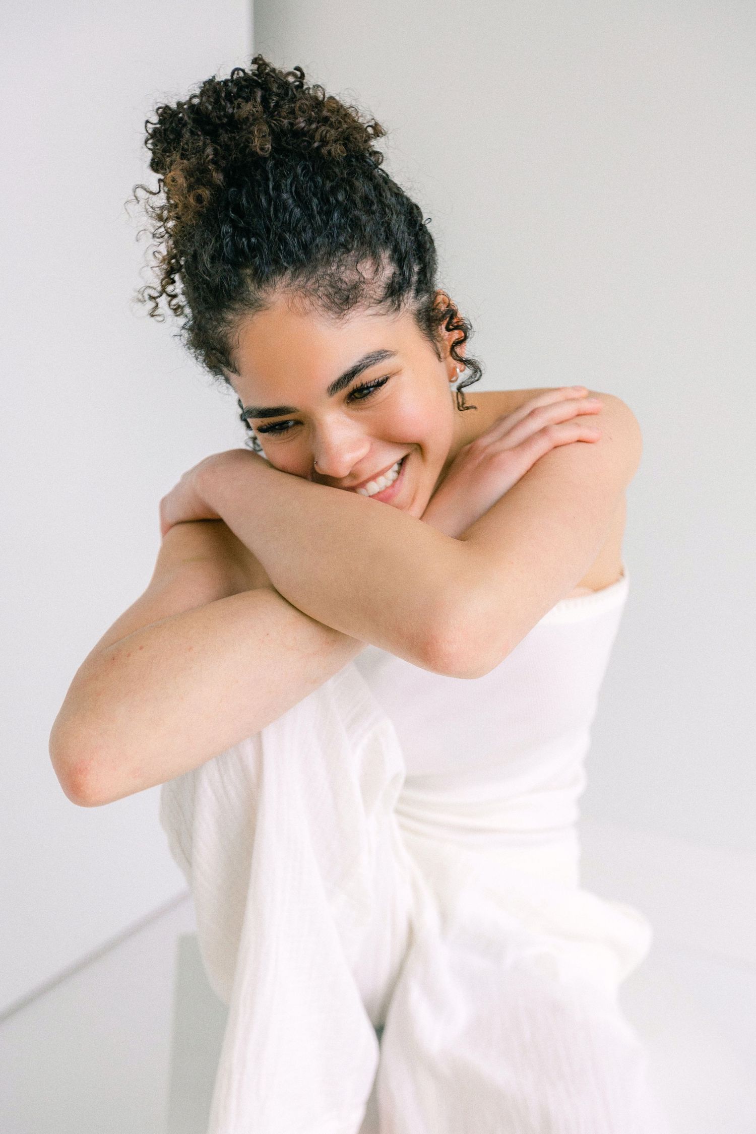Smiling young woman with curly updo hair, wearing white, arms crossed over chest against light background.