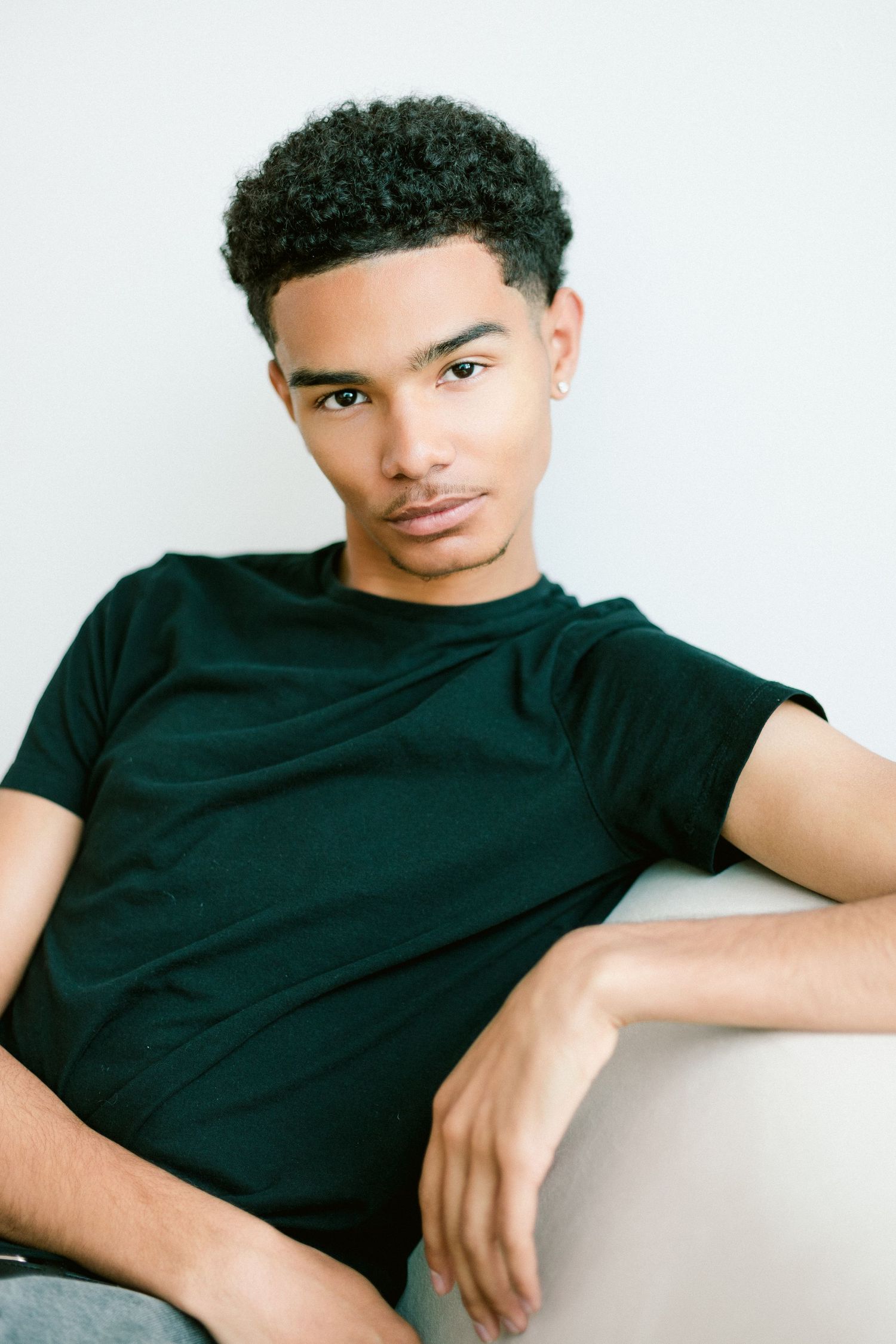 Young Black man in black t-shirt sitting relaxed against white wall, looking at camera with confident expression.