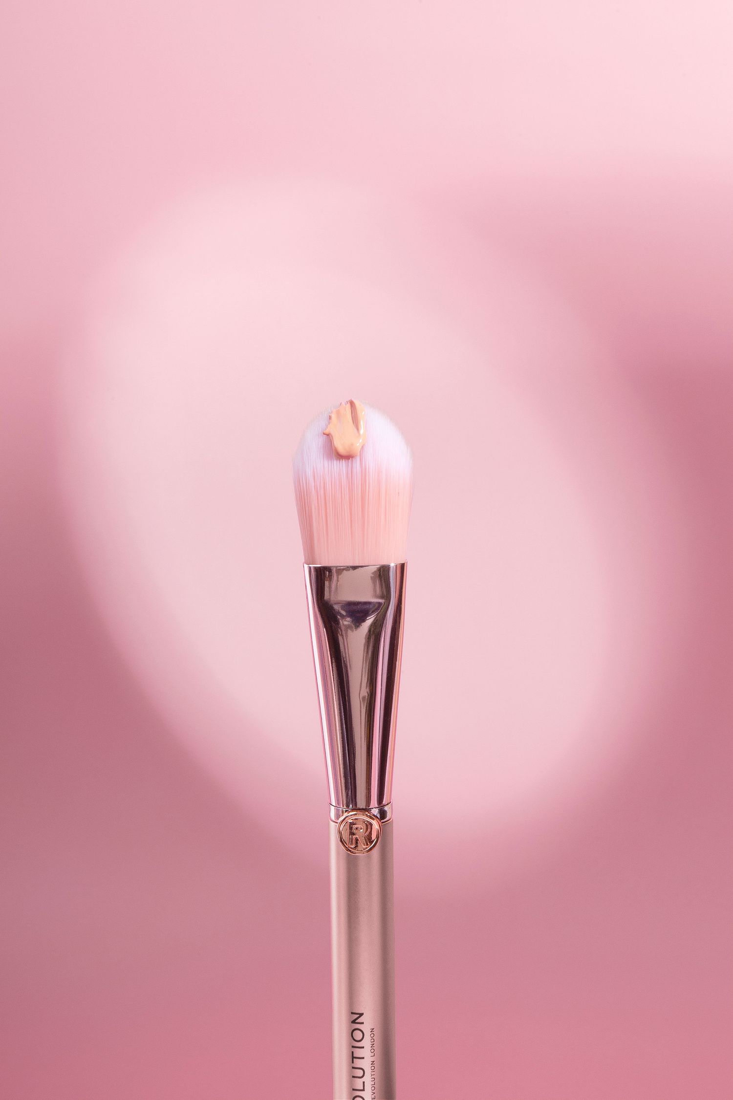 Pink makeup brush with foundation drop on bristles against soft pink background, rose gold handle beauty tool.