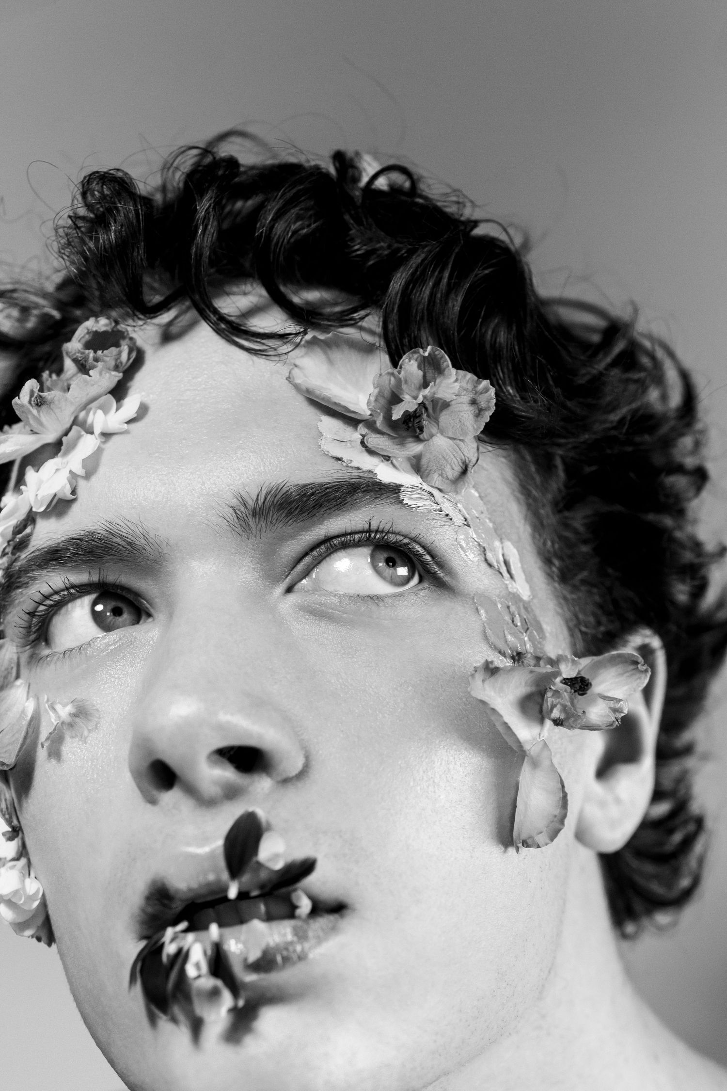 Black and white portrait of a young man with curly hair and flowers adorning his face, gazing upward.