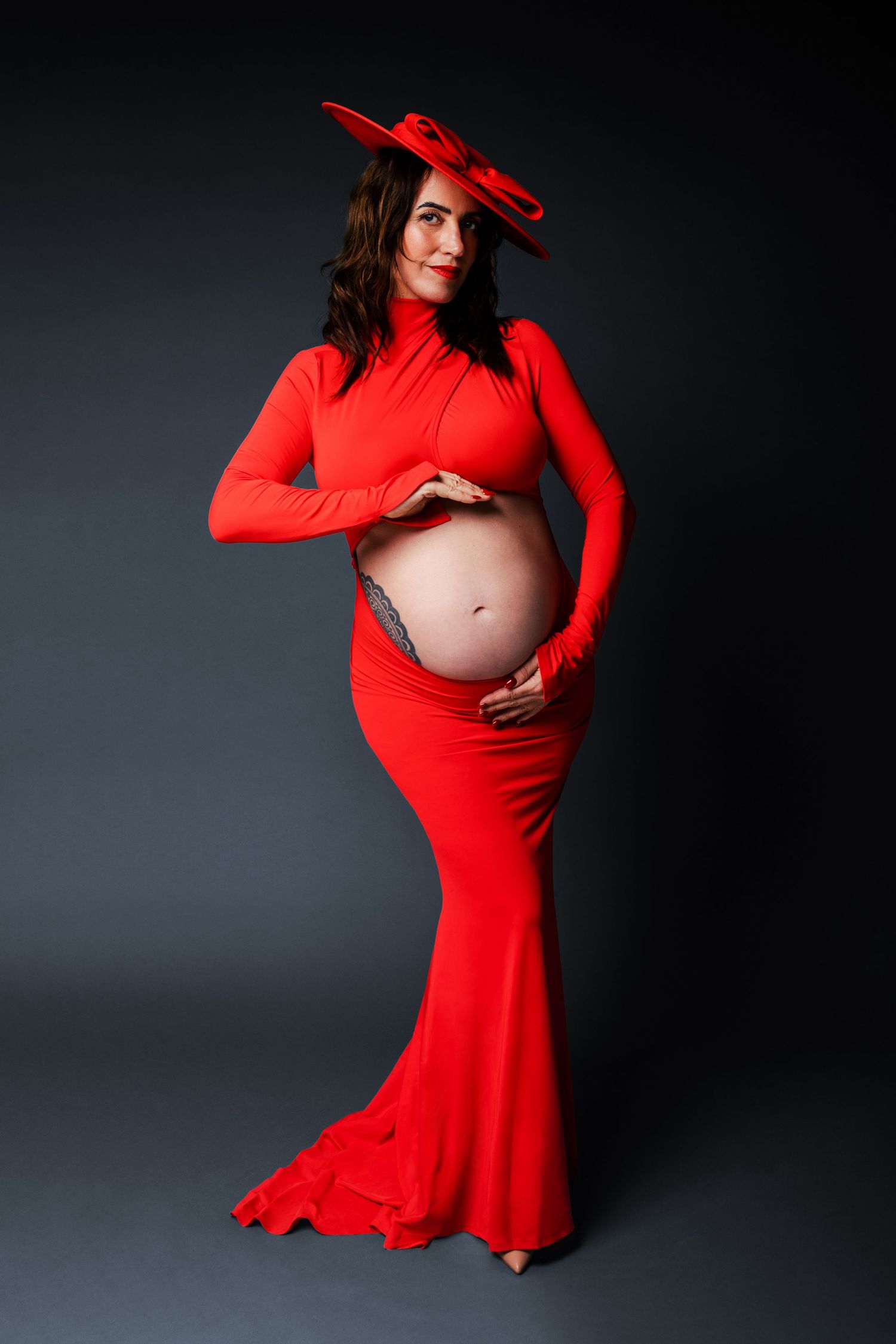 Pregnant woman in elegant red gown and hat, cradling baby bump, posing against dark studio background.