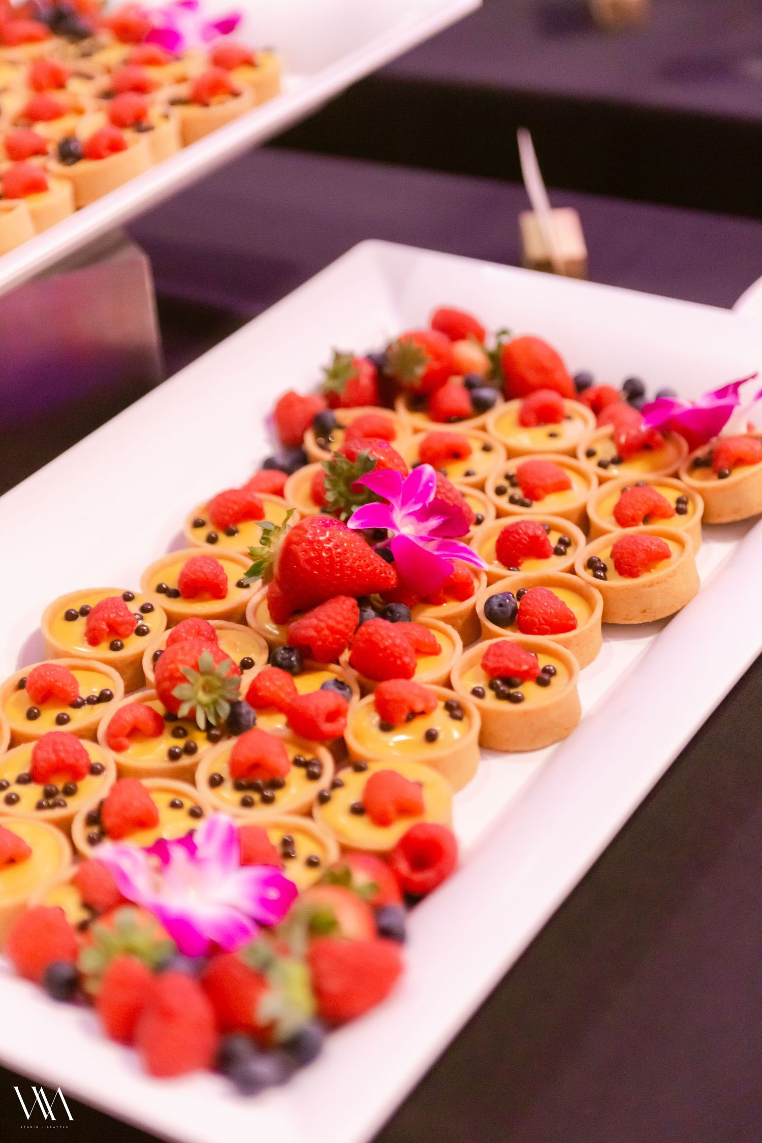 Elegant dessert tray with fresh fruit tarts topped with strawberries, raspberries, blueberries, and pink orchid flowers.