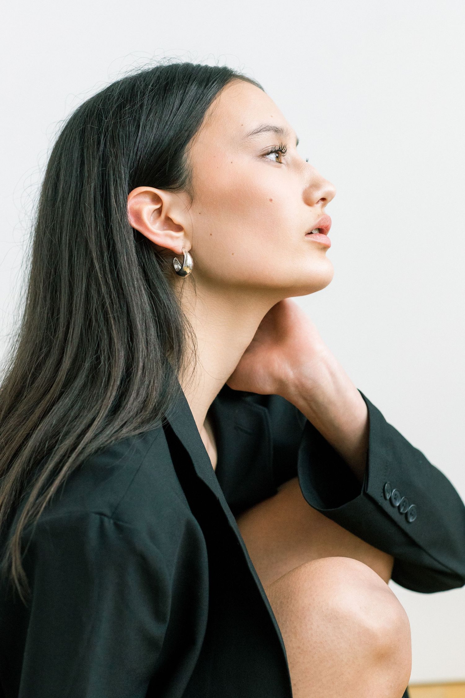 A woman with long dark hair in a black blazer, hand on neck, gazing upward, wearing gold earrings.
