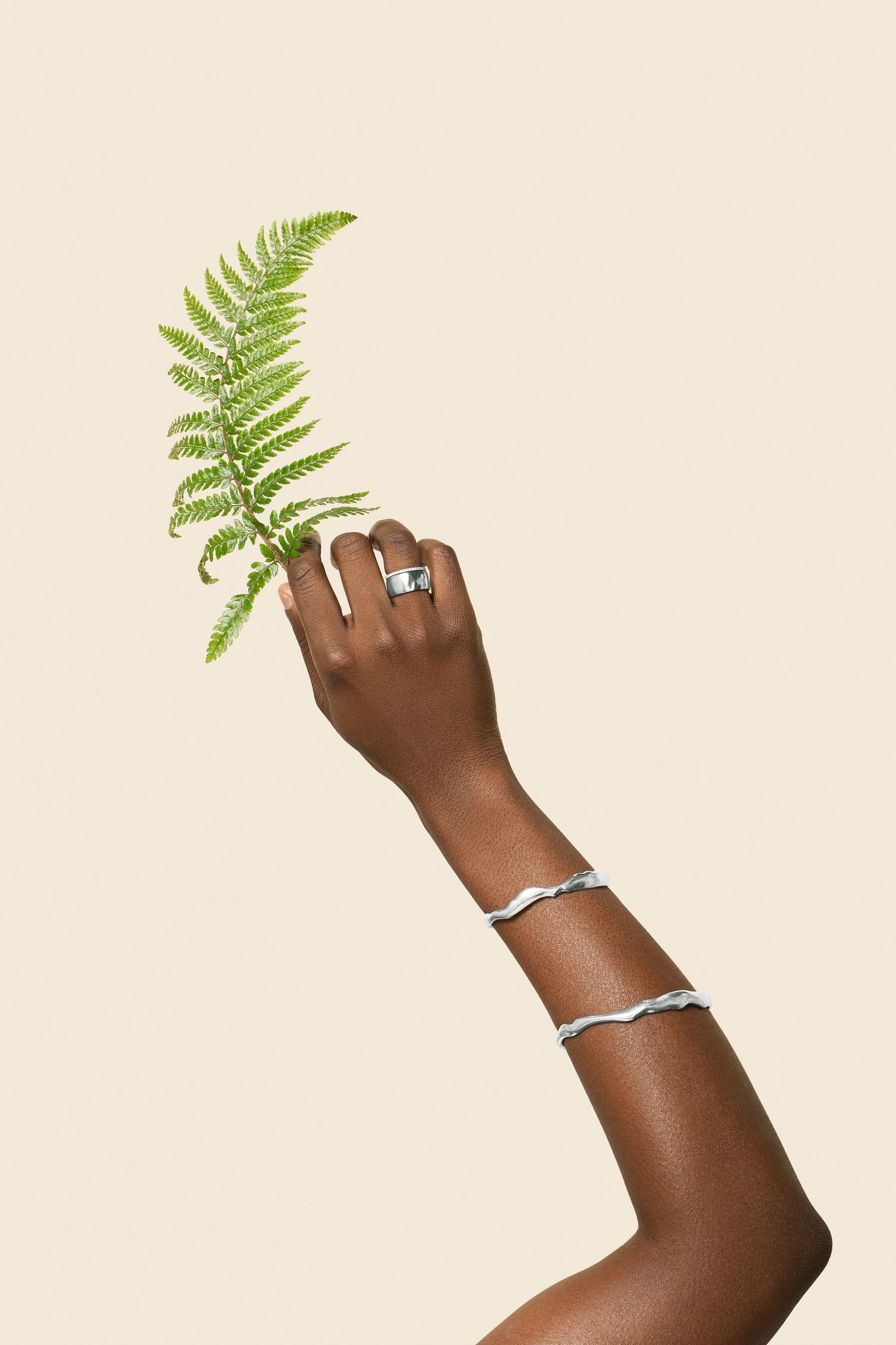 Dark-skinned hand holding a green fern leaf, adorned with a silver ring and two bracelets, against a beige background.