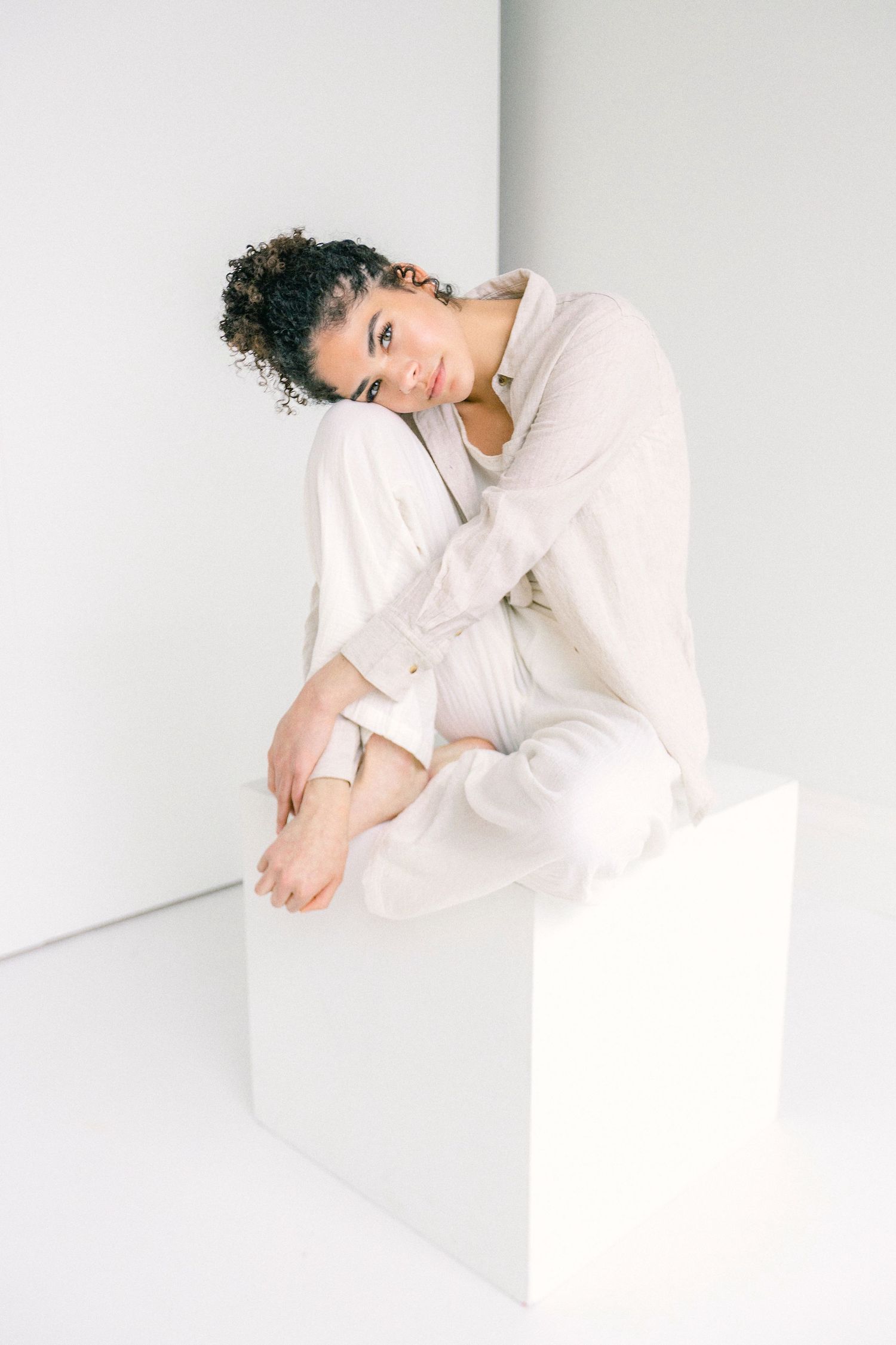 A young woman with curly hair sits barefoot on a white cube, wearing a beige linen outfit against a white background.