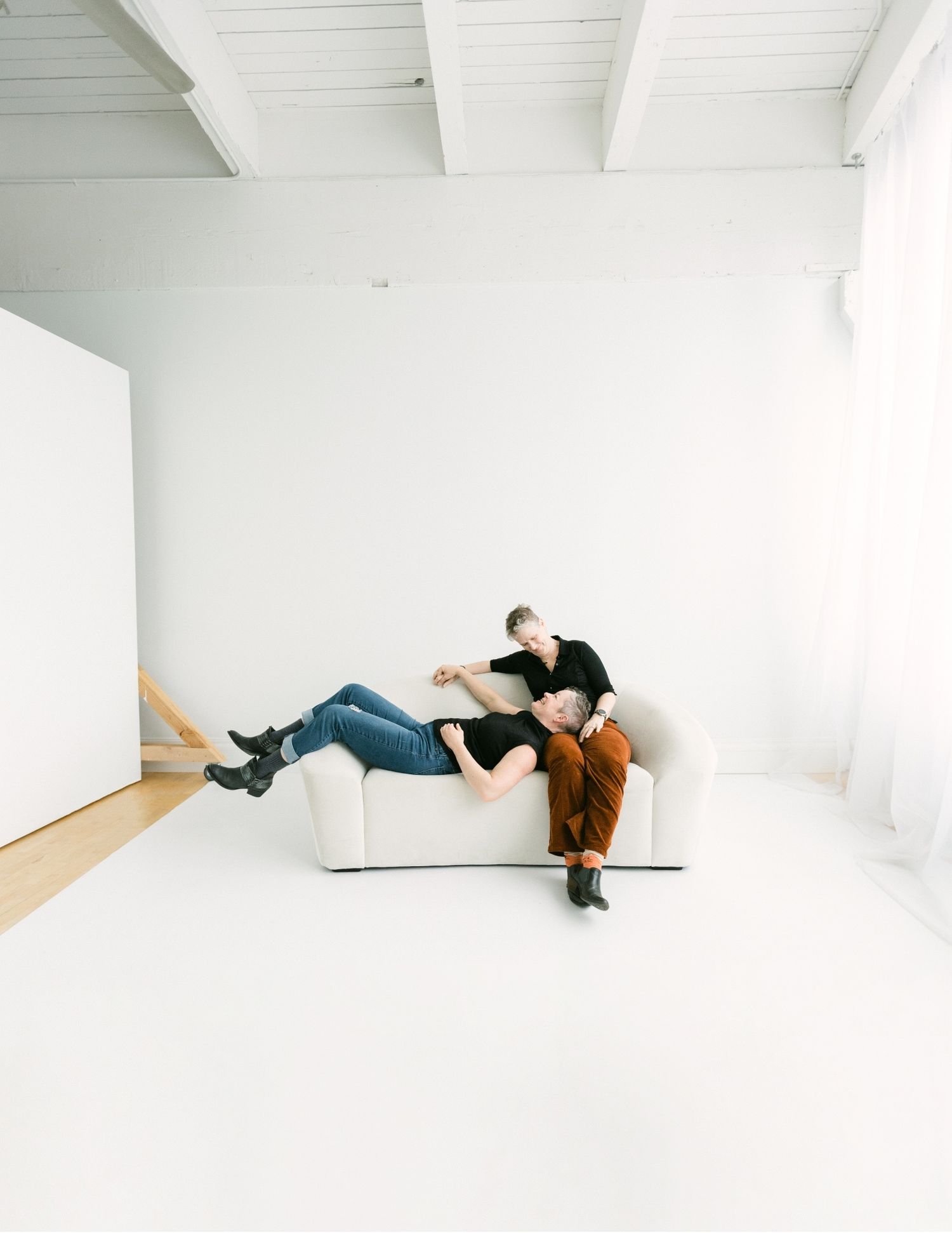 Two women lounging playfully on a white sofa in a bright open studio with white floors and ceiling.