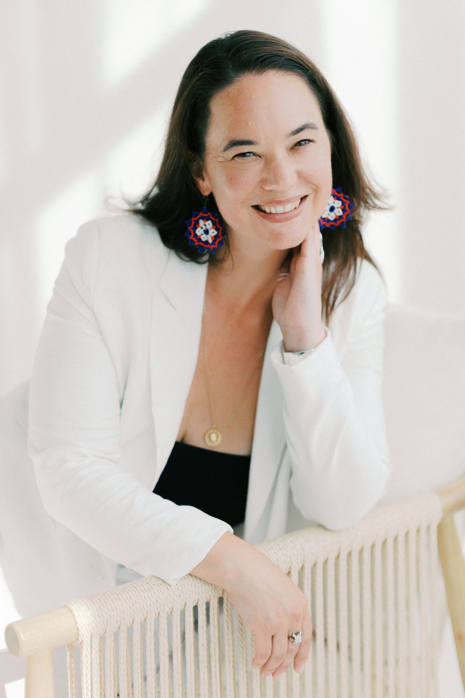 Smiling woman in white blazer with colorful floral earrings, resting chin on hand while seated in a bright room.