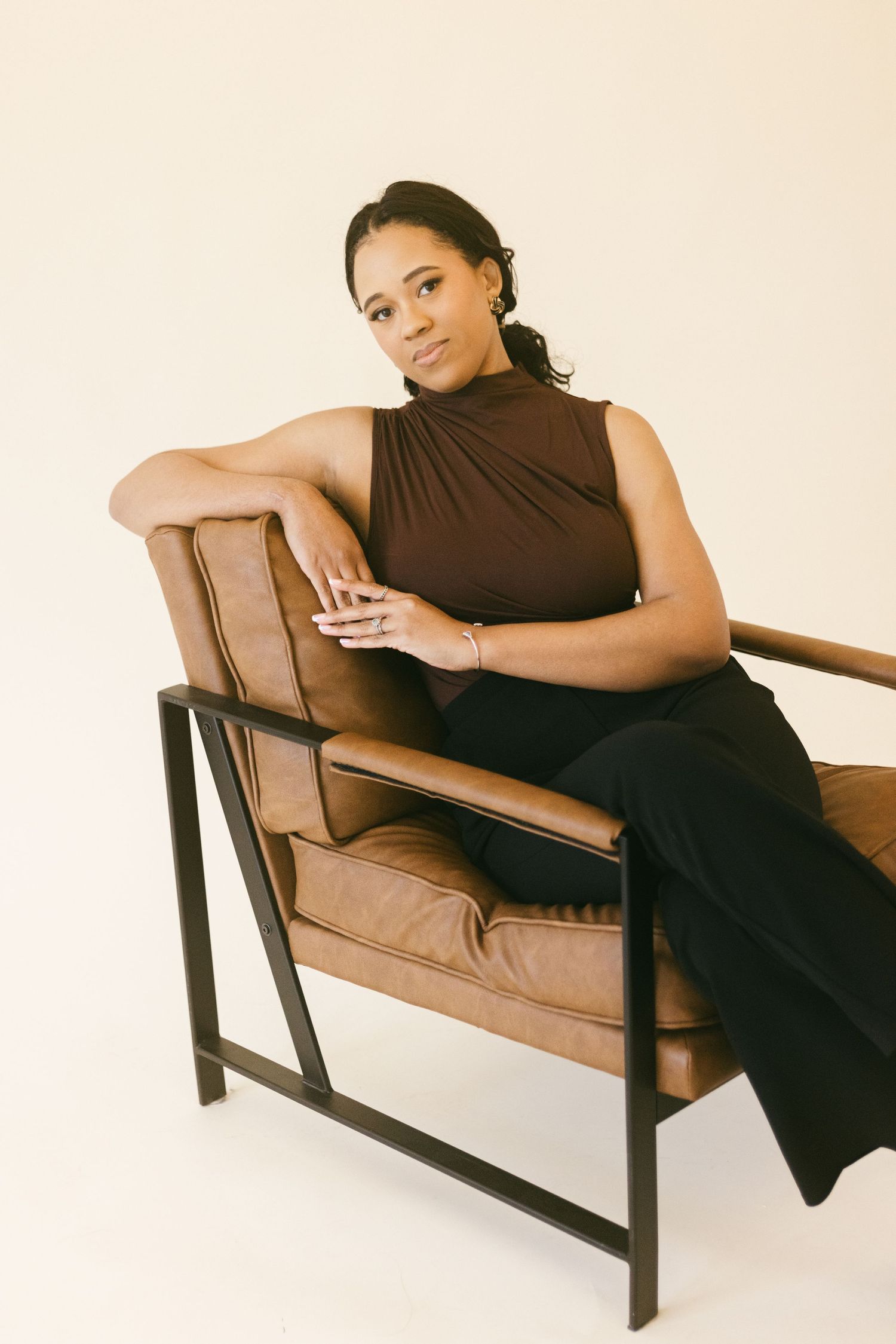 Woman in brown sleeveless top and black pants seated elegantly in a brown leather chair against a neutral background.