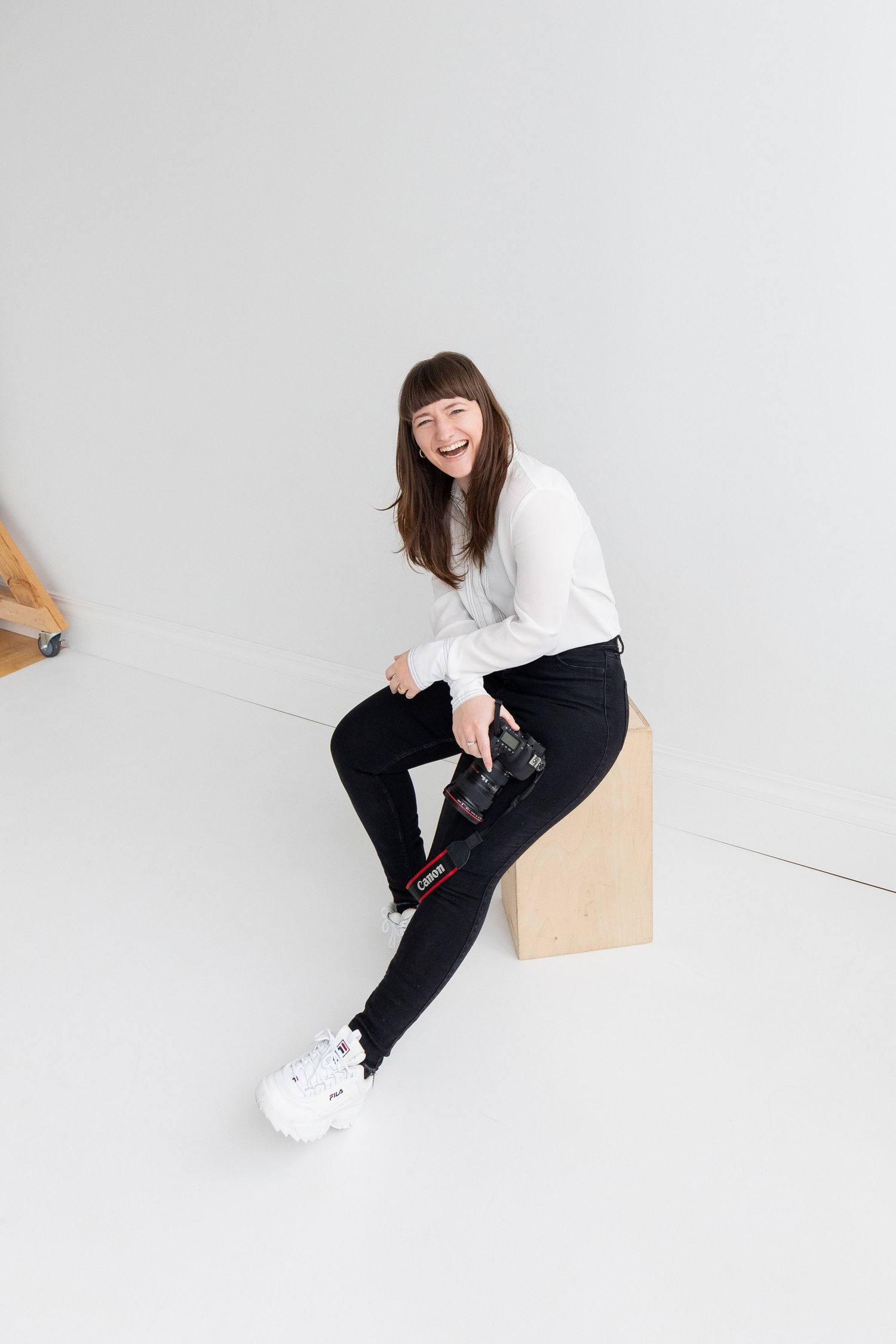 A laughing female photographer sits on a wooden cube holding a camera in a clean white studio space.