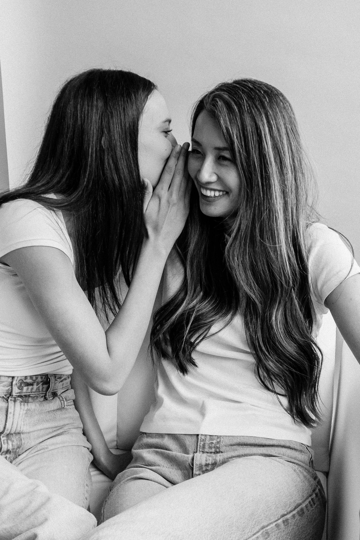 Mother and daughter share a secret, one whispering to the other who laughs in black and white.