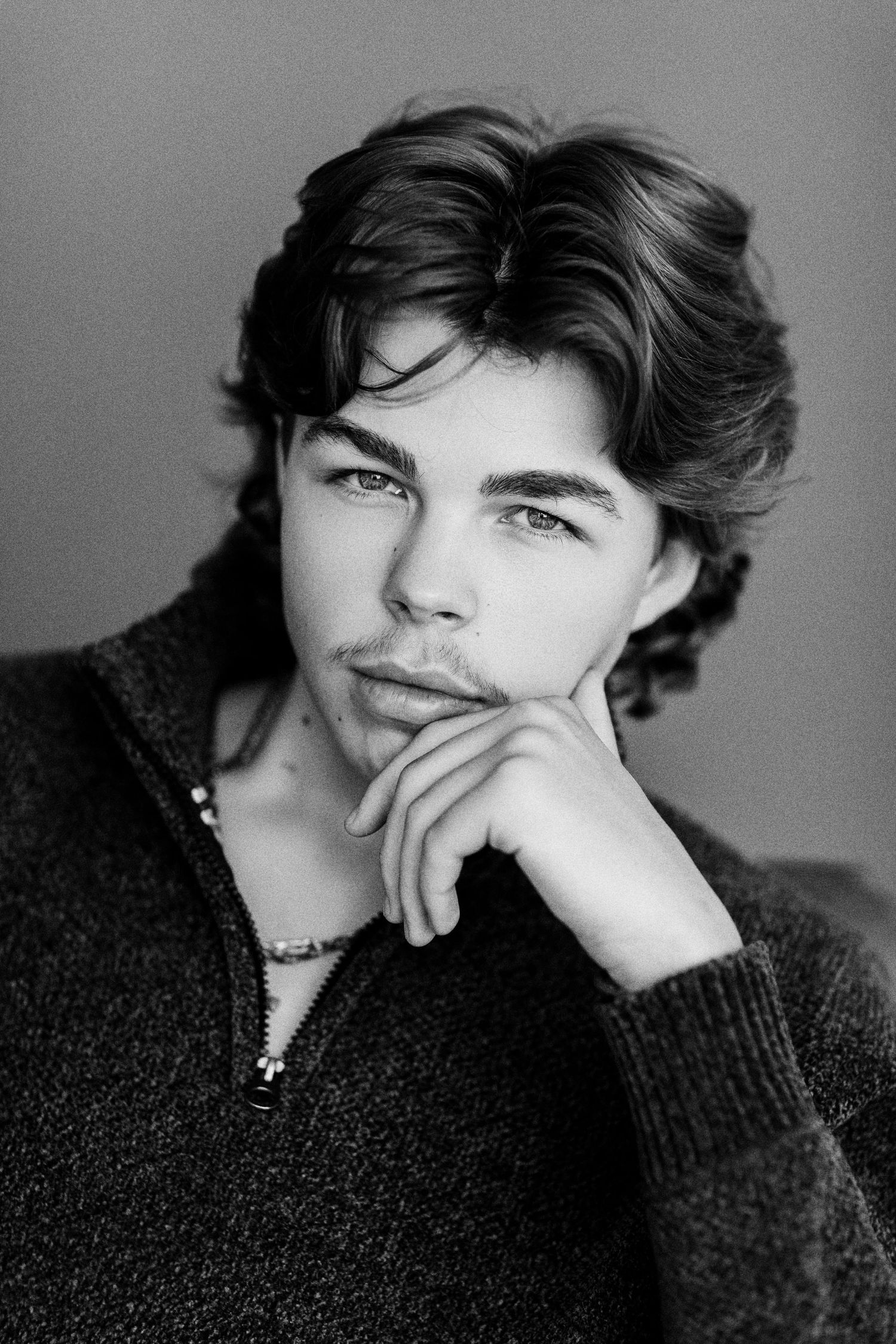Black and white portrait of a young man with wavy hair resting chin on hand, wearing a knit sweater and necklace.