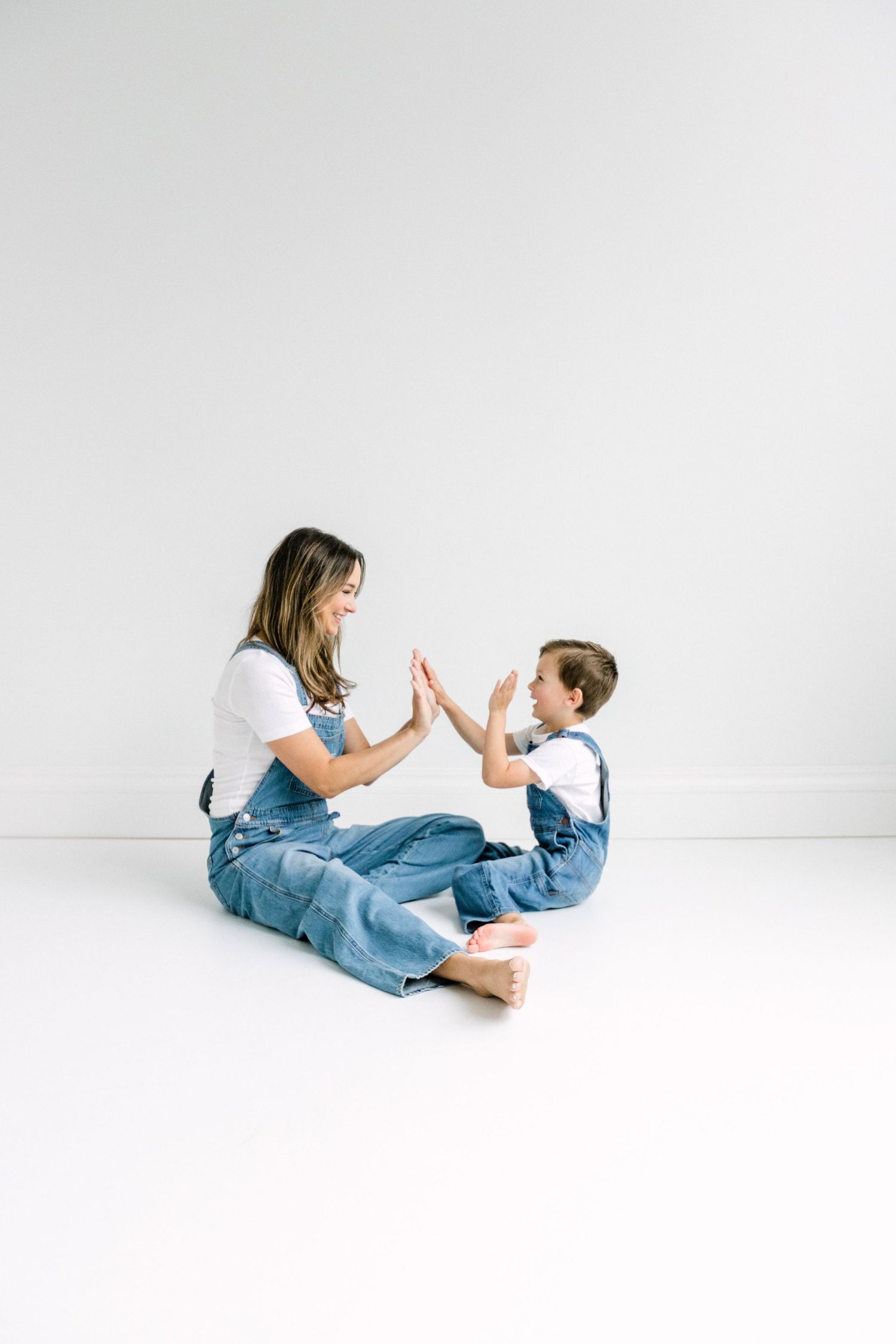 A mother and child in matching denim overalls sit on a white floor playing a clapping game, smiling at each other.