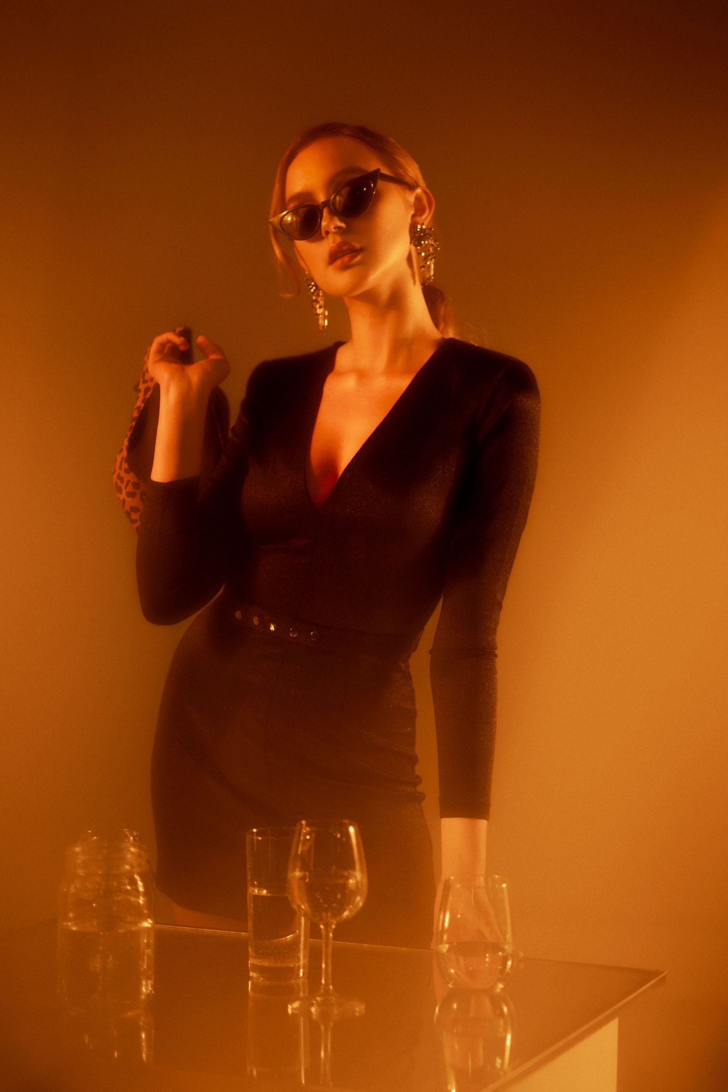 A woman in a black dress and sunglasses poses elegantly by a glass table under warm orange lighting.