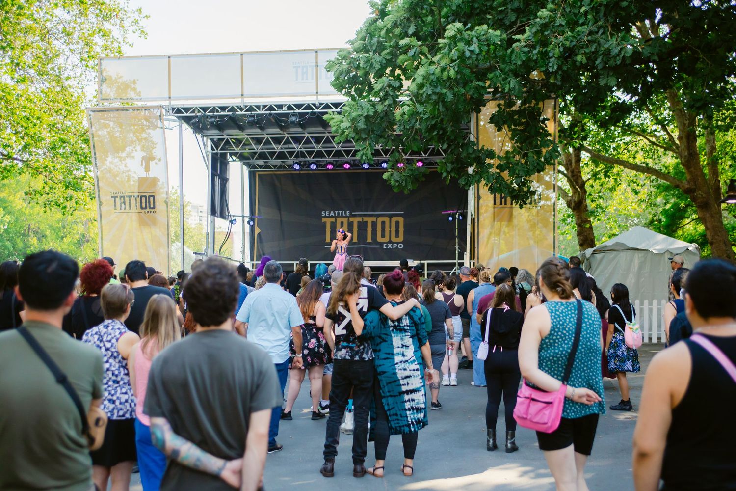 Crowd gathered at an outdoor Tattoo festival stage surrounded by lush green trees on a sunny day.