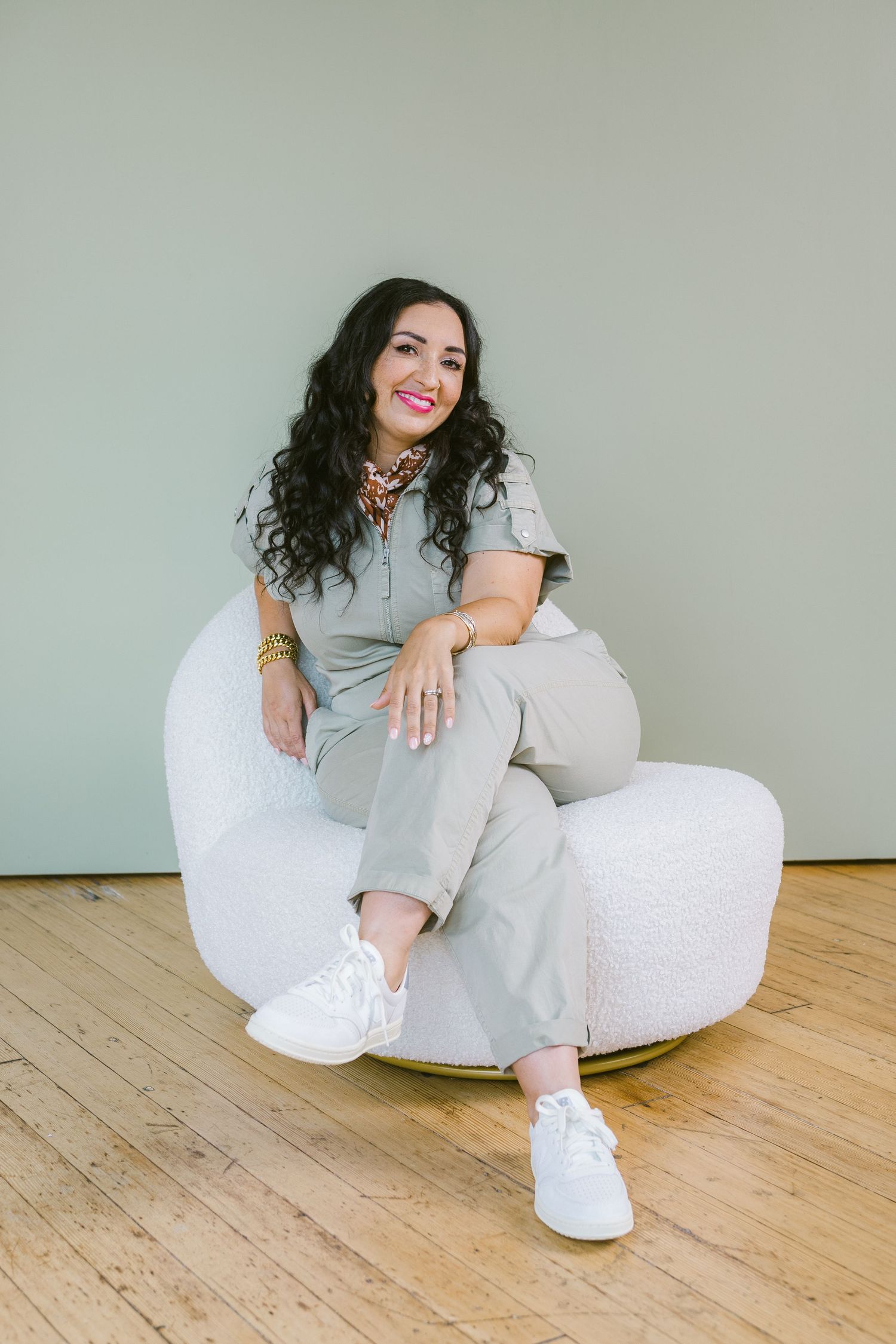 Woman in sage green jumpsuit sitting in white chair, smiling, with curly dark hair and white sneakers.