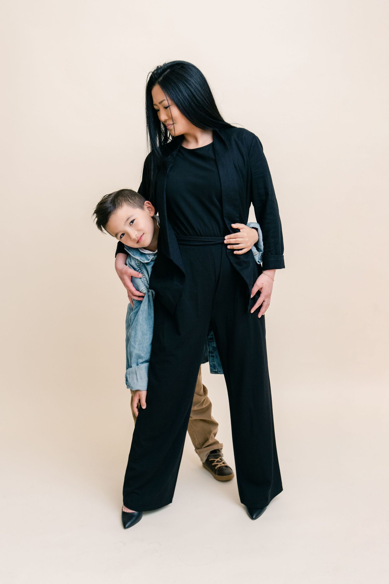 A smiling young boy hugs his stylish mother dressed in a black pantsuit against a beige background.