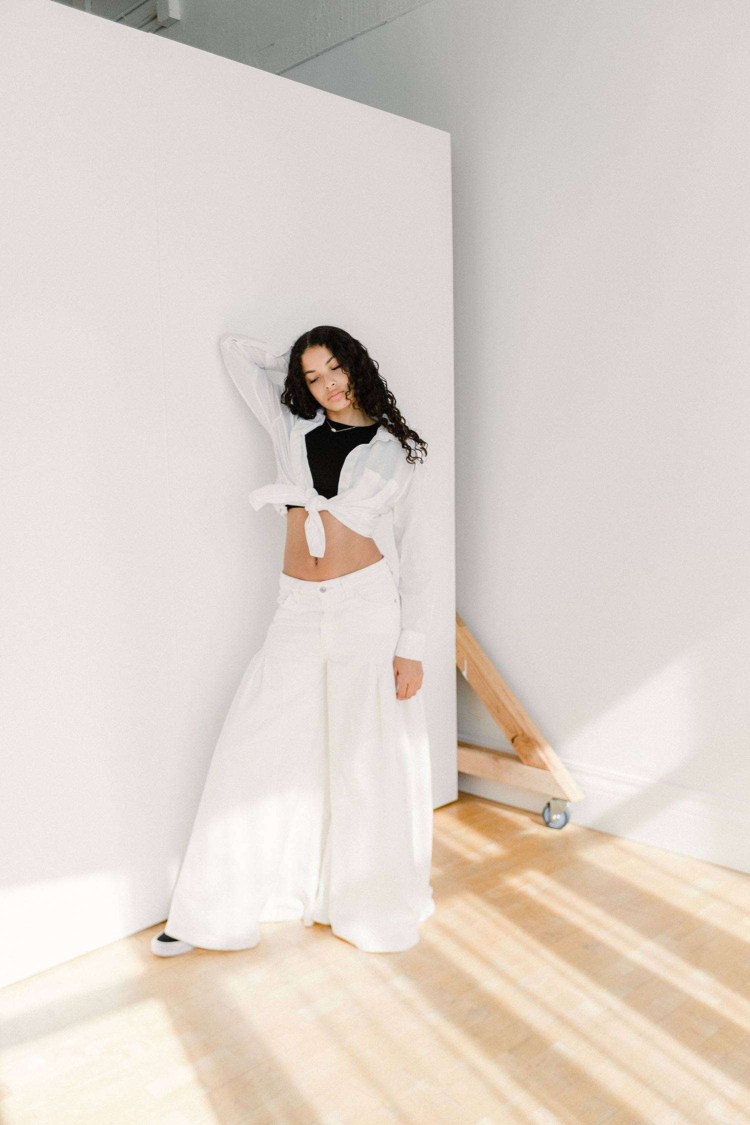 Young woman in white wide-leg pants and black crop top posing in bright white studio.