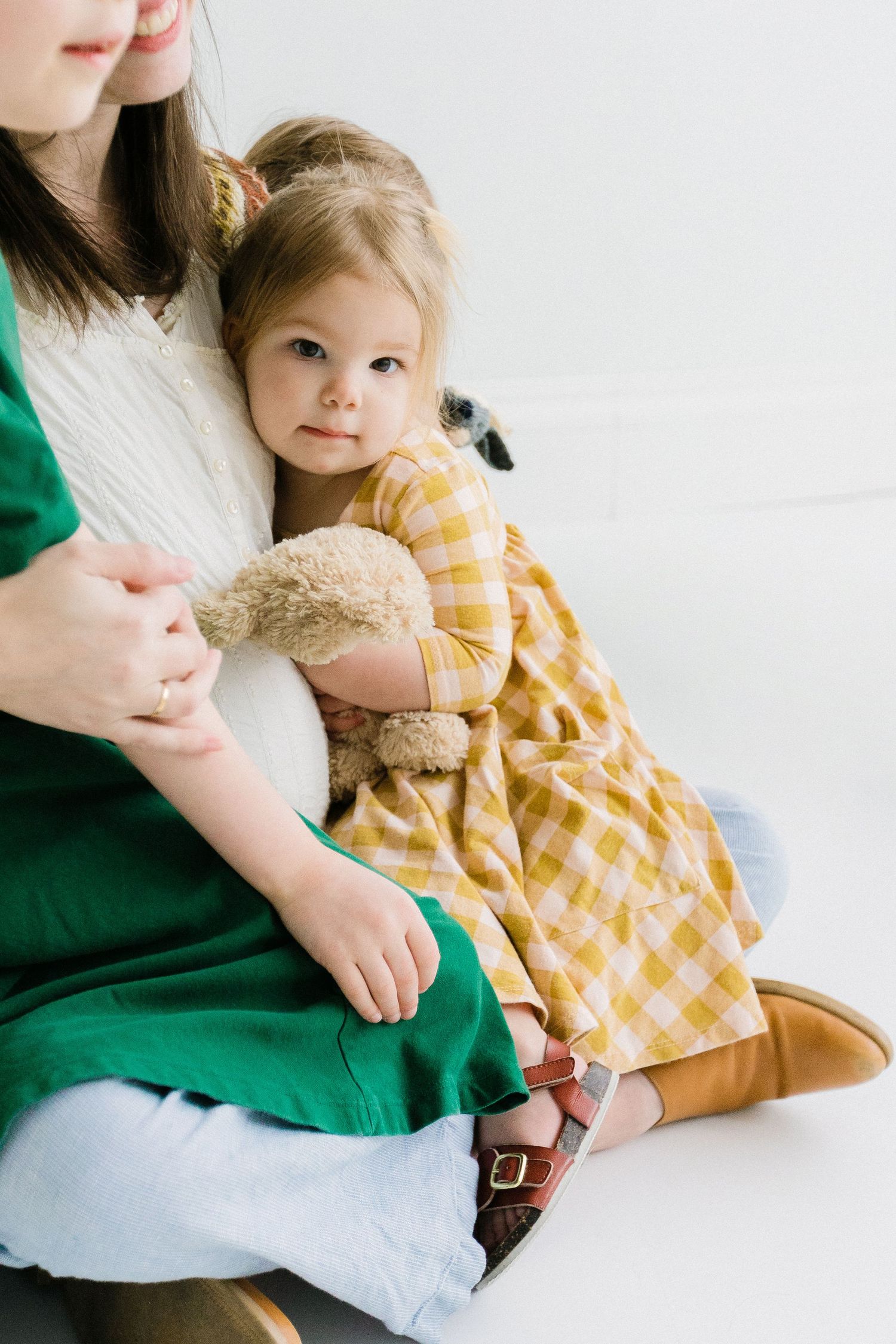 Young toddler girl in yellow gingham dress holding stuffed animal sitting with mother.