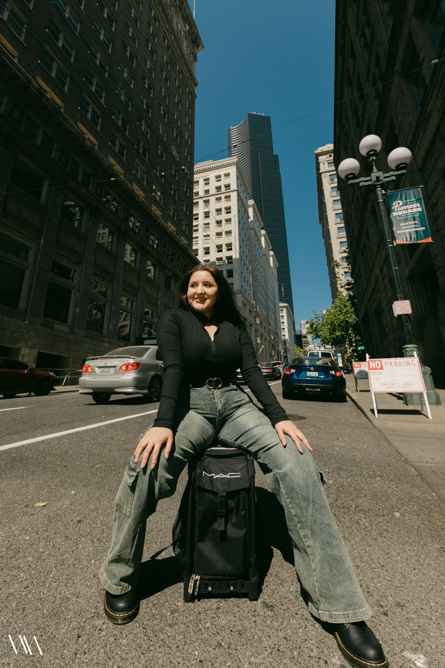 Woman in black top and jeans sits on MAC luggage in urban street surrounded by tall city buildings on sunny day.