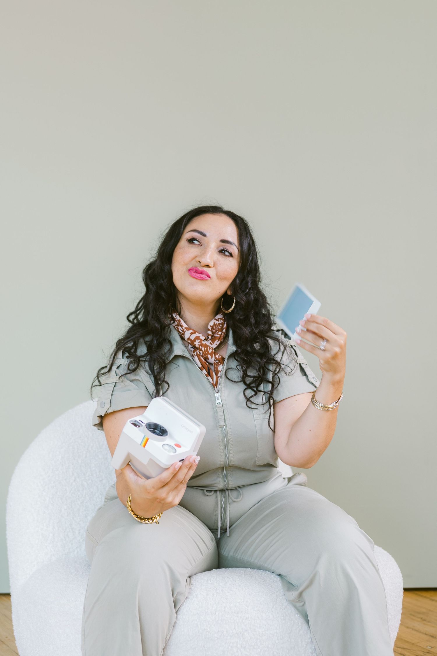 Woman in gray jumpsuit sitting in white chair holding an instant camera and photo print.