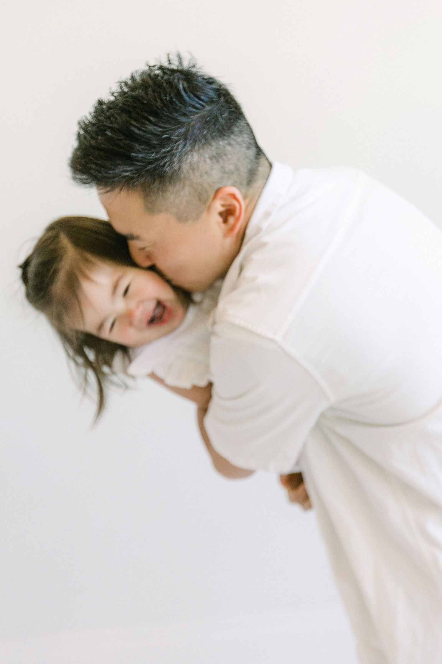A father kisses his laughing child while carrying them in his arms against a white background.