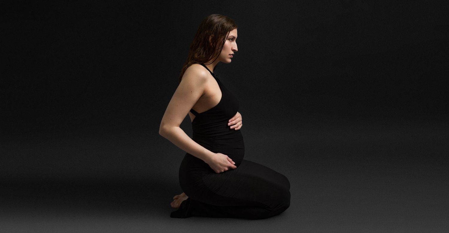 Pregnant woman in black outfit sitting gracefully on floor against dark background, cradling baby bump.