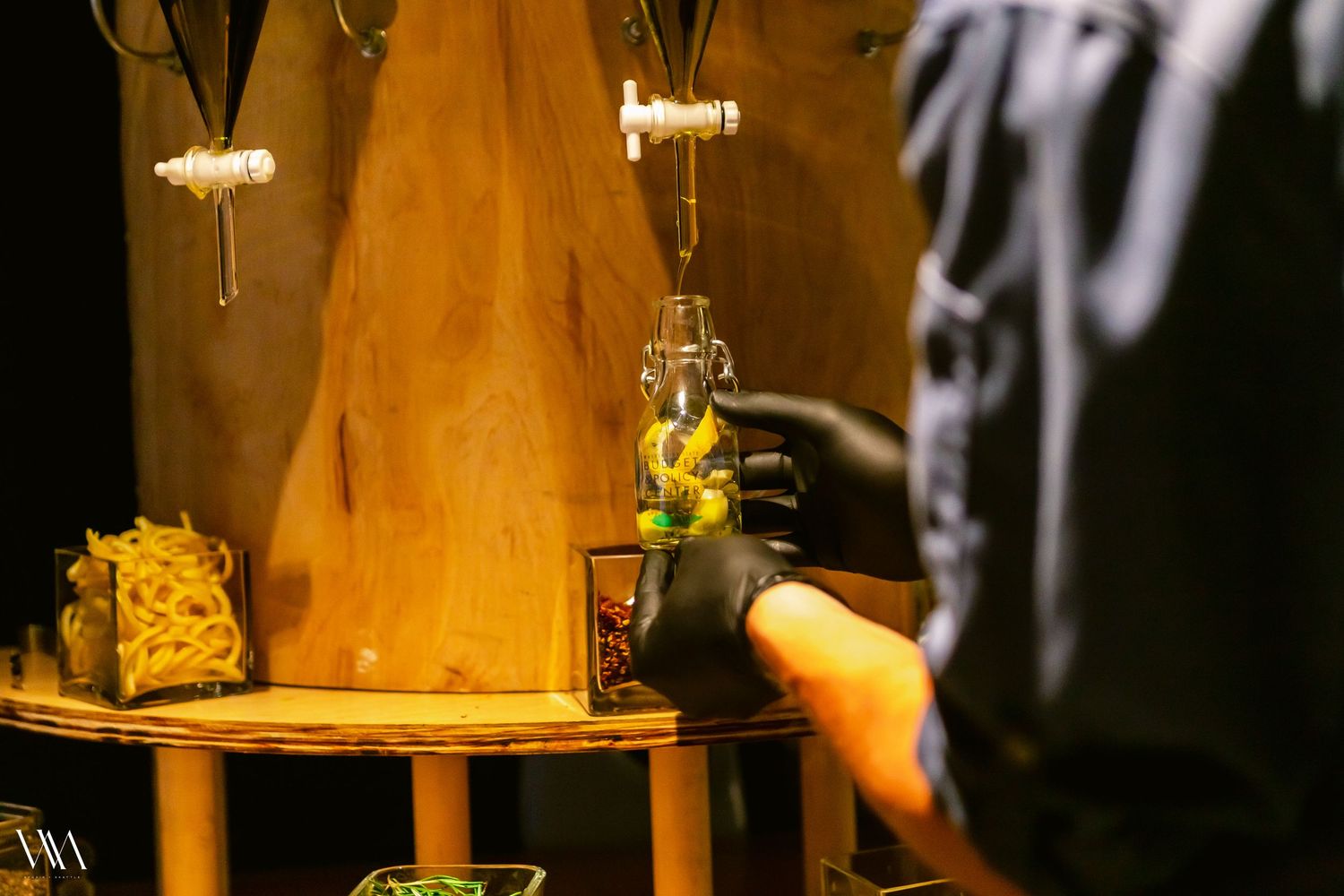 A bartender in black gloves pours a drink into a glass bottle on a wooden bar display with taps.