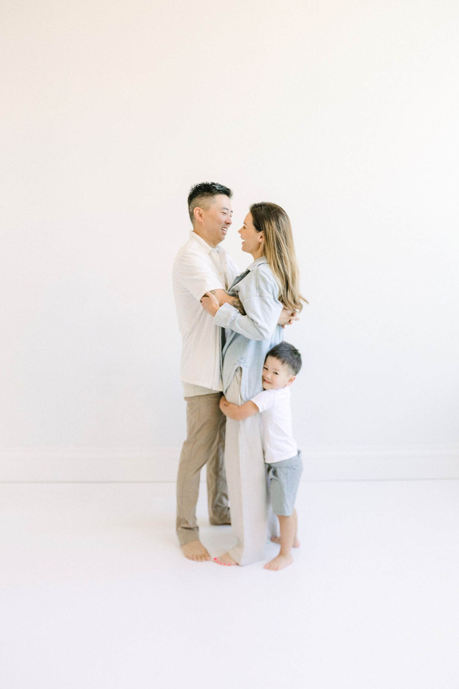 A happy Asian family of three poses together in a bright white studio, with parents embracing and toddler hugging mom's legs.