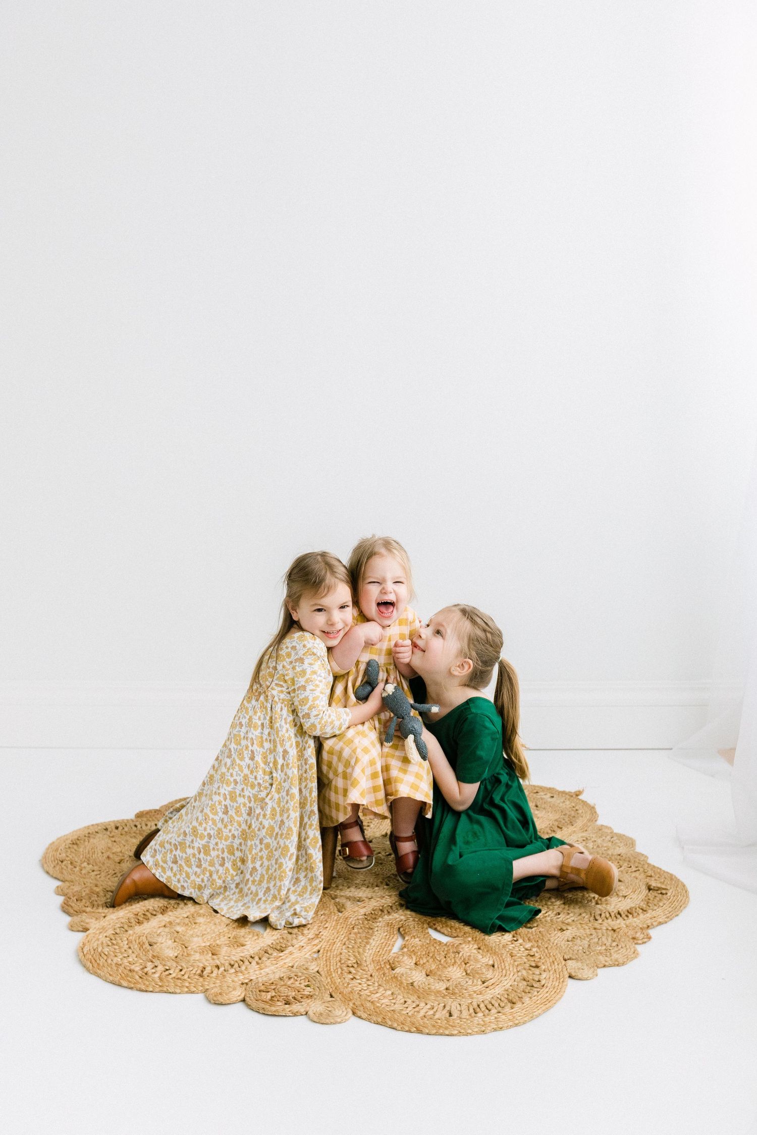 Three young girls in colorful dresses laughing and playing together on a woven rug against a white wall.