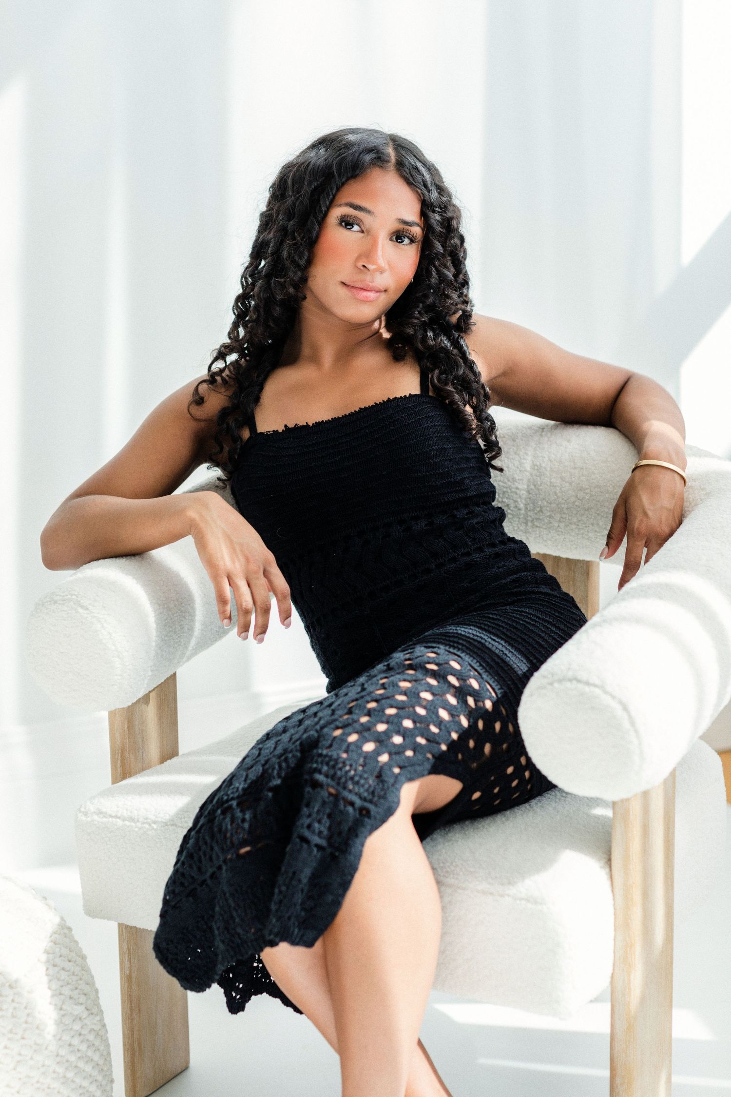 Young woman with curly black hair sits gracefully in a white chair, wearing a black crochet outfit against a white backdrop.