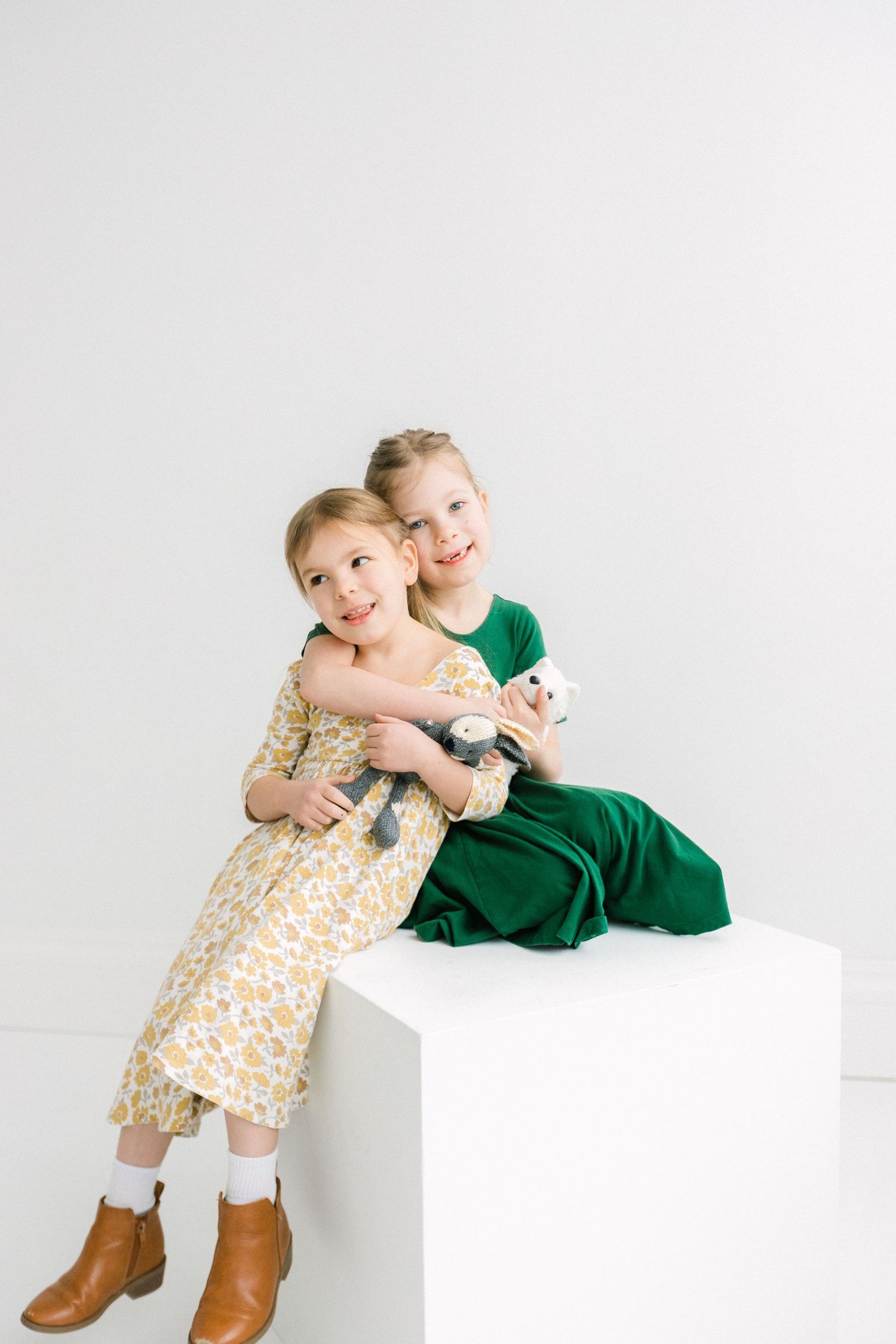 Two little girls in colorful dresses sit together on a white cube holding stuffed animals.