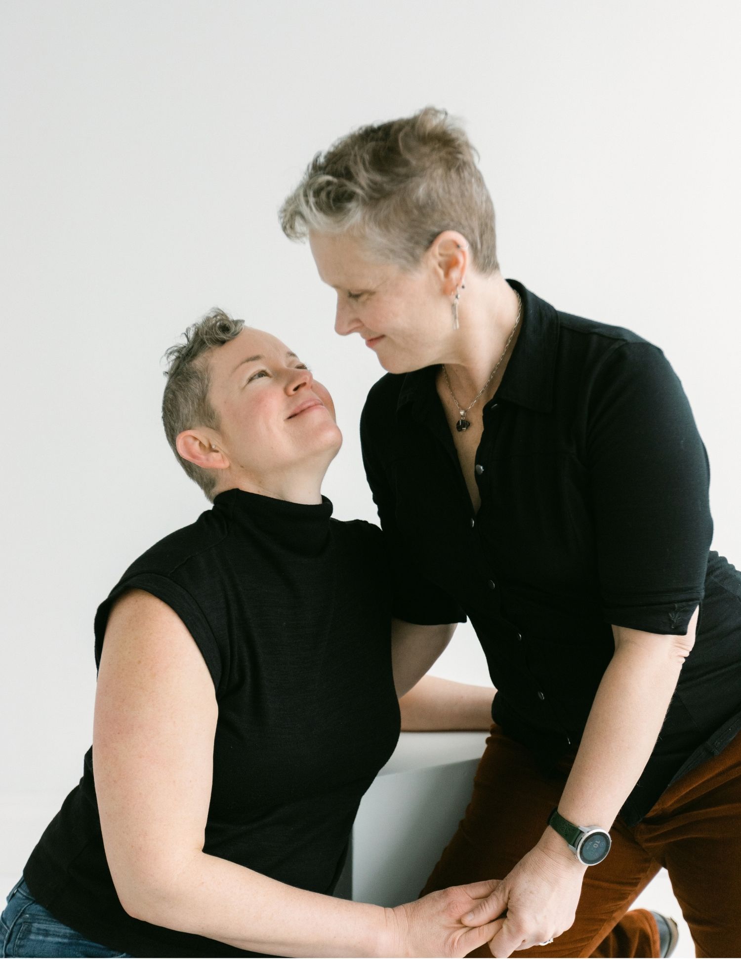 Two people in black tops share a tender moment, gazing at each other affectionately against a white background.
