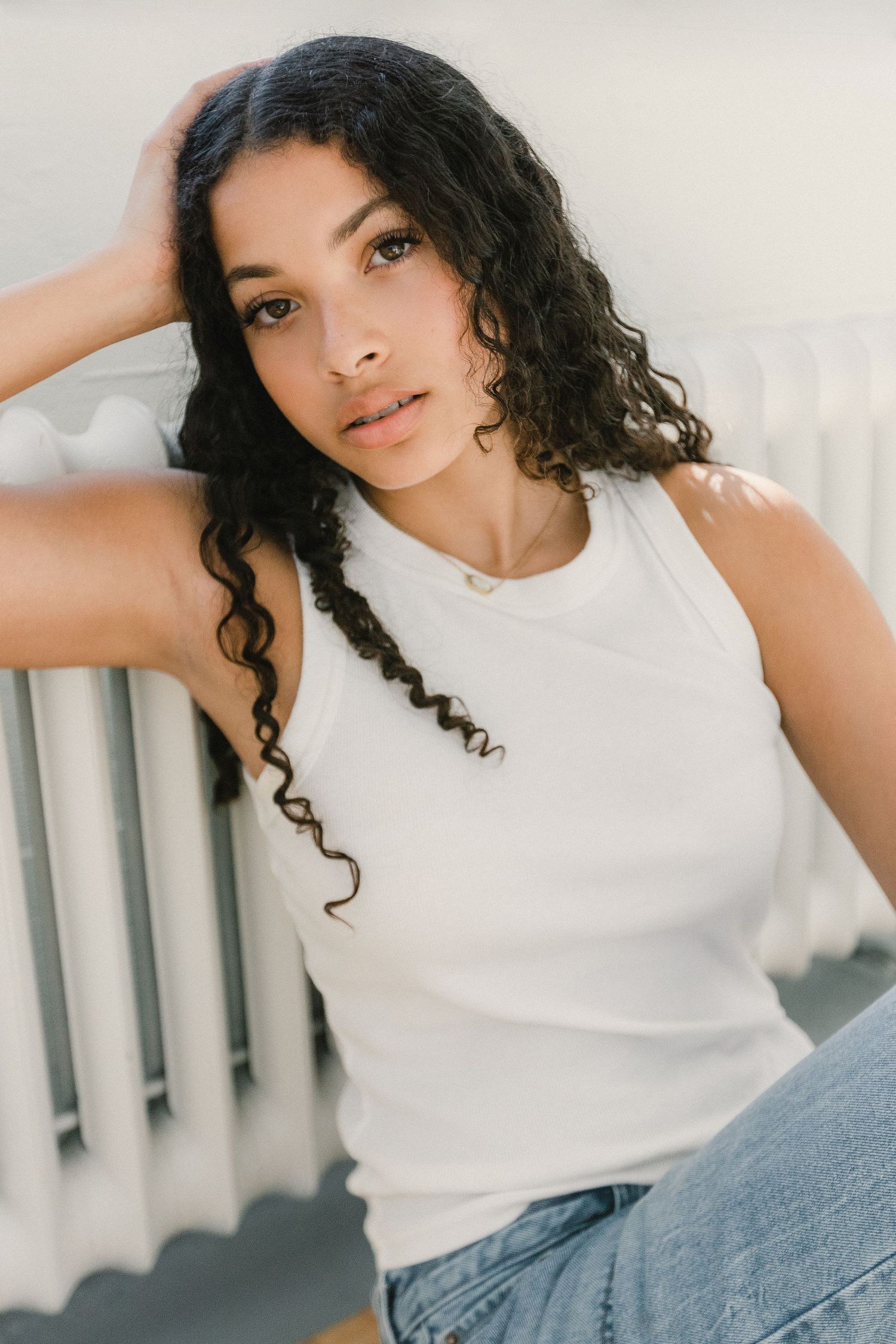 Young woman with curly dark hair in white tank top leaning against radiator in sunlight.