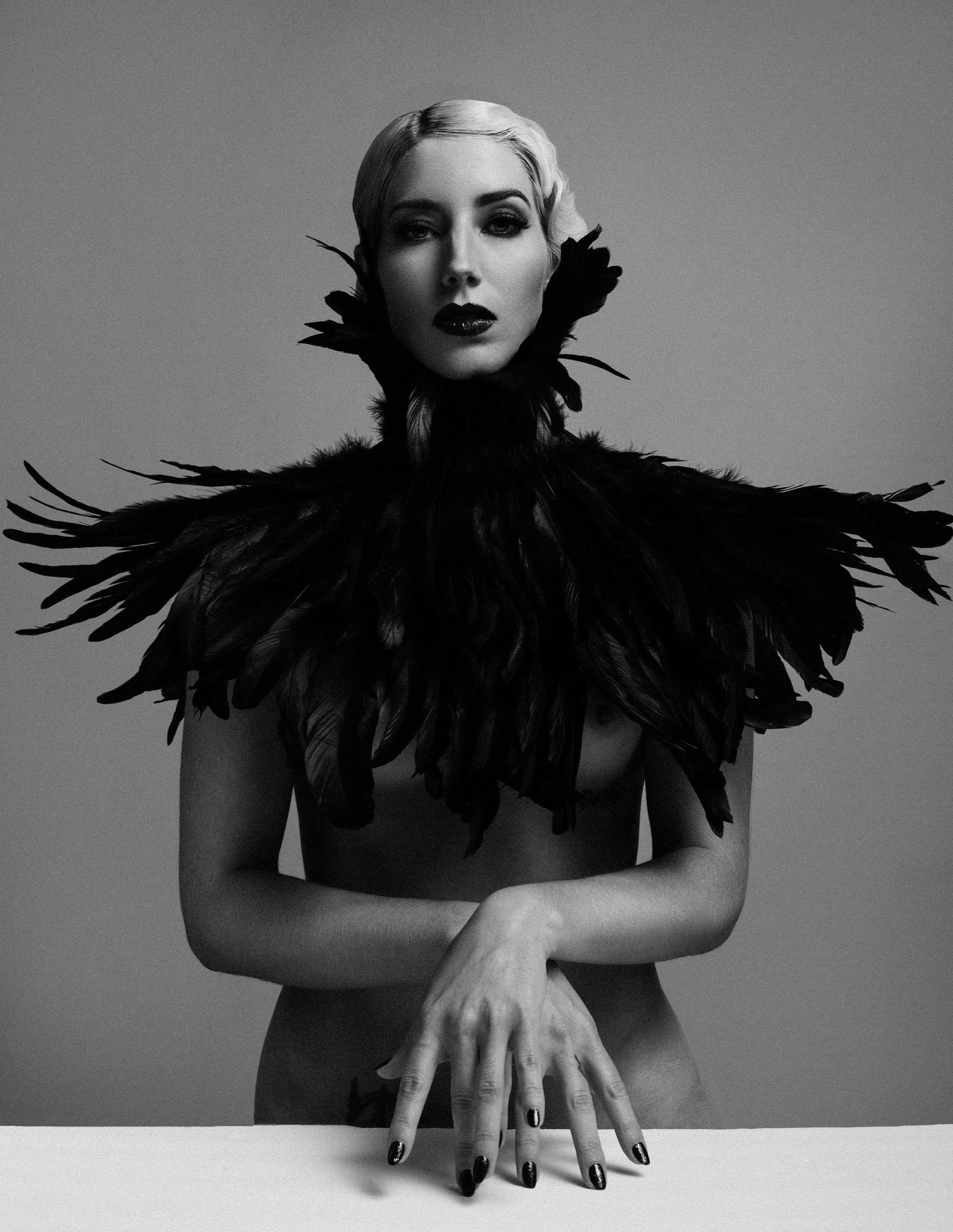 Black and white portrait of a woman wearing dramatic black feather shoulder piece, arms crossed on white surface.