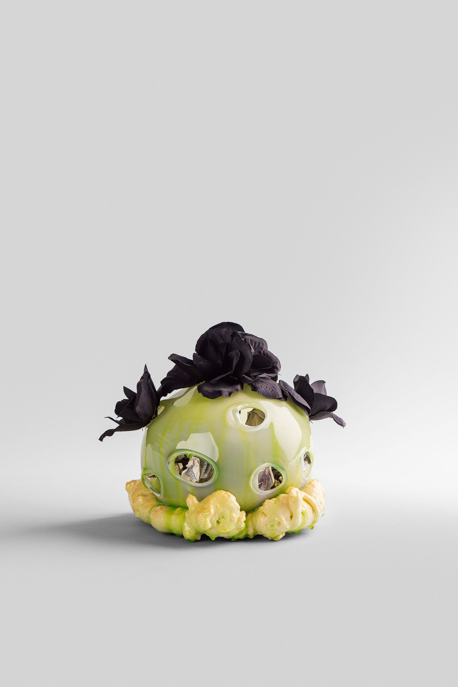 Green ceramic sculpture with black flower details and yellow base, featuring decorative holes, on white background.