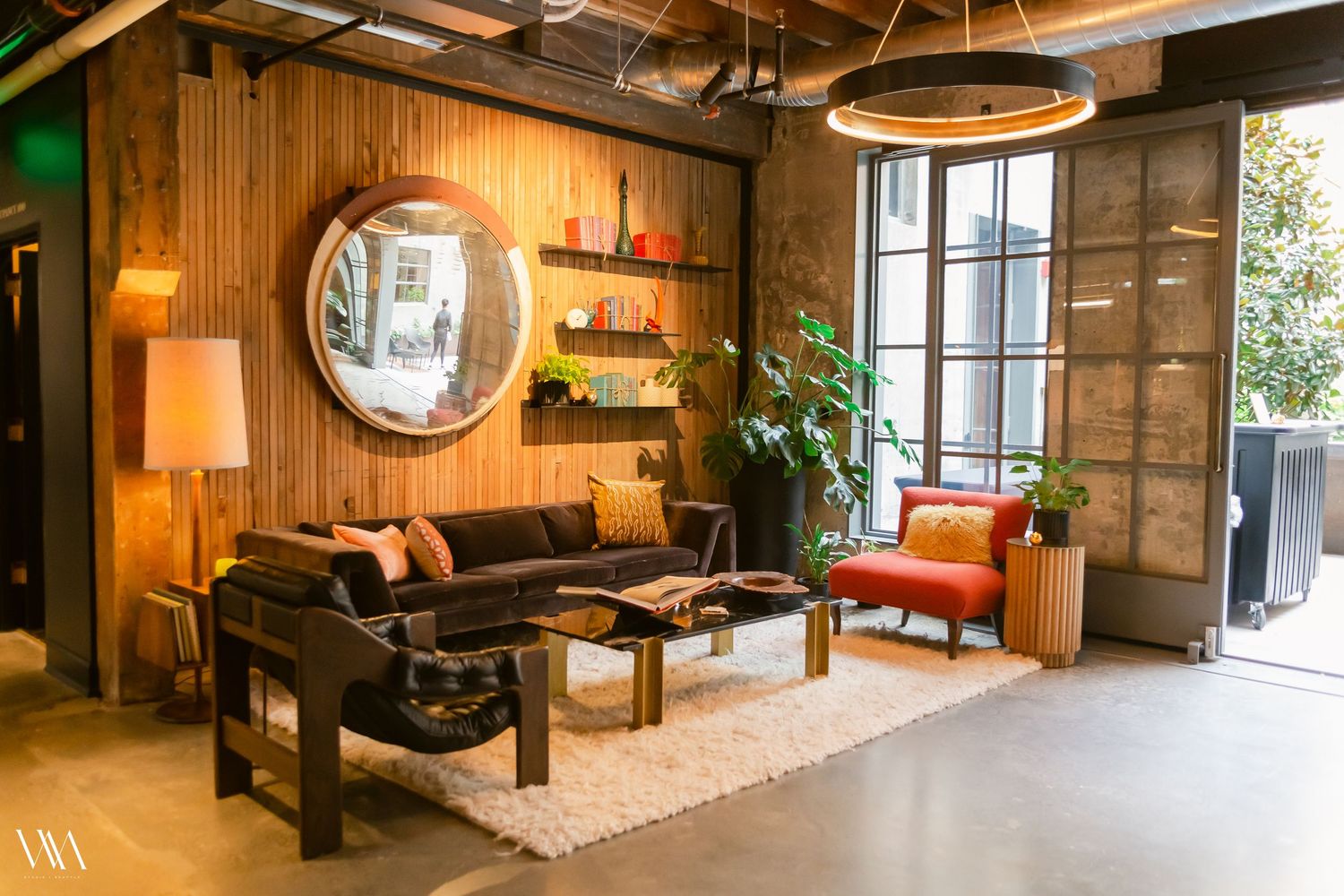 Cozy mid-century modern living room with wood-paneled walls, round mirror, dark sofa, red accent chair, and lush plants.