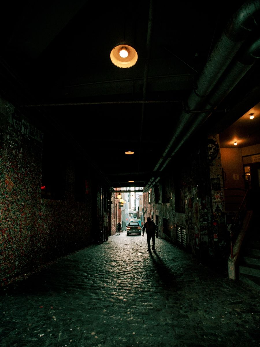 Dark cobblestone alley with glowing pendant light illuminating silhouette walking through Seattle gum wall tunnel.