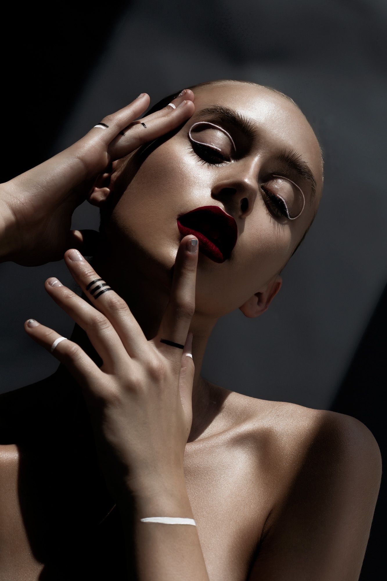 Dramatic beauty portrait of bald model with dark red lips and multiple rings posing with hands near face.