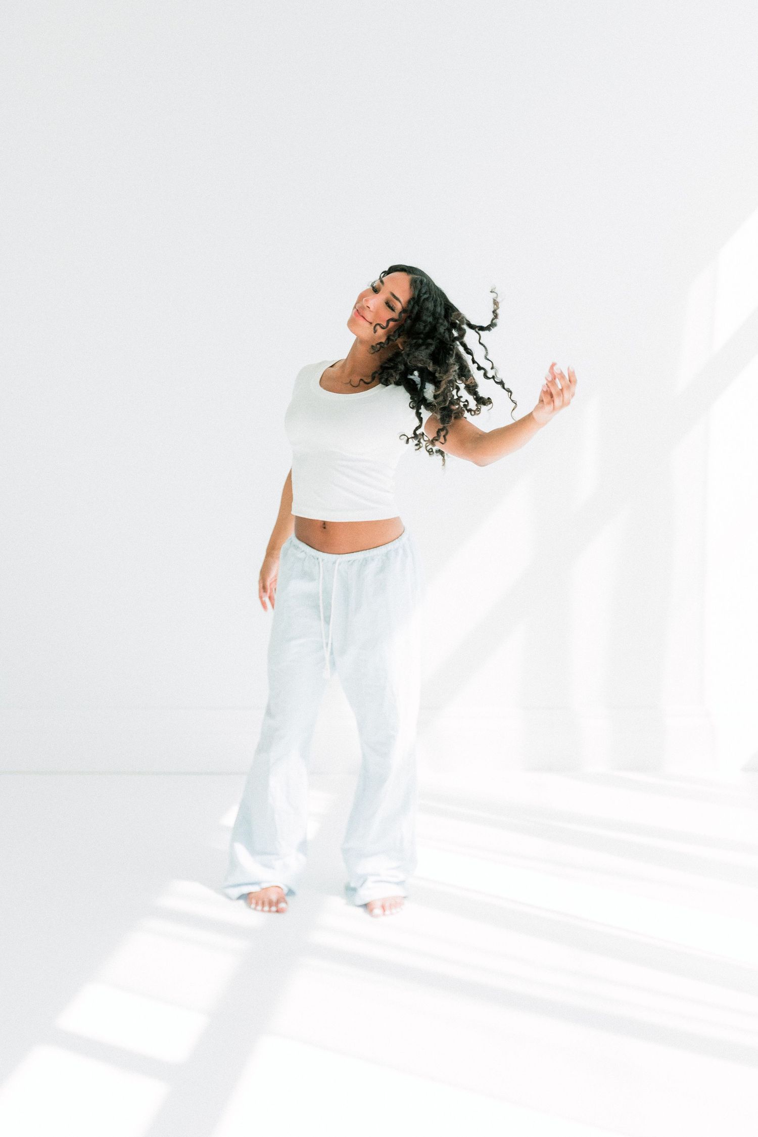 Woman with curly hair dancing in white crop top and light blue flare pants against a bright white background.