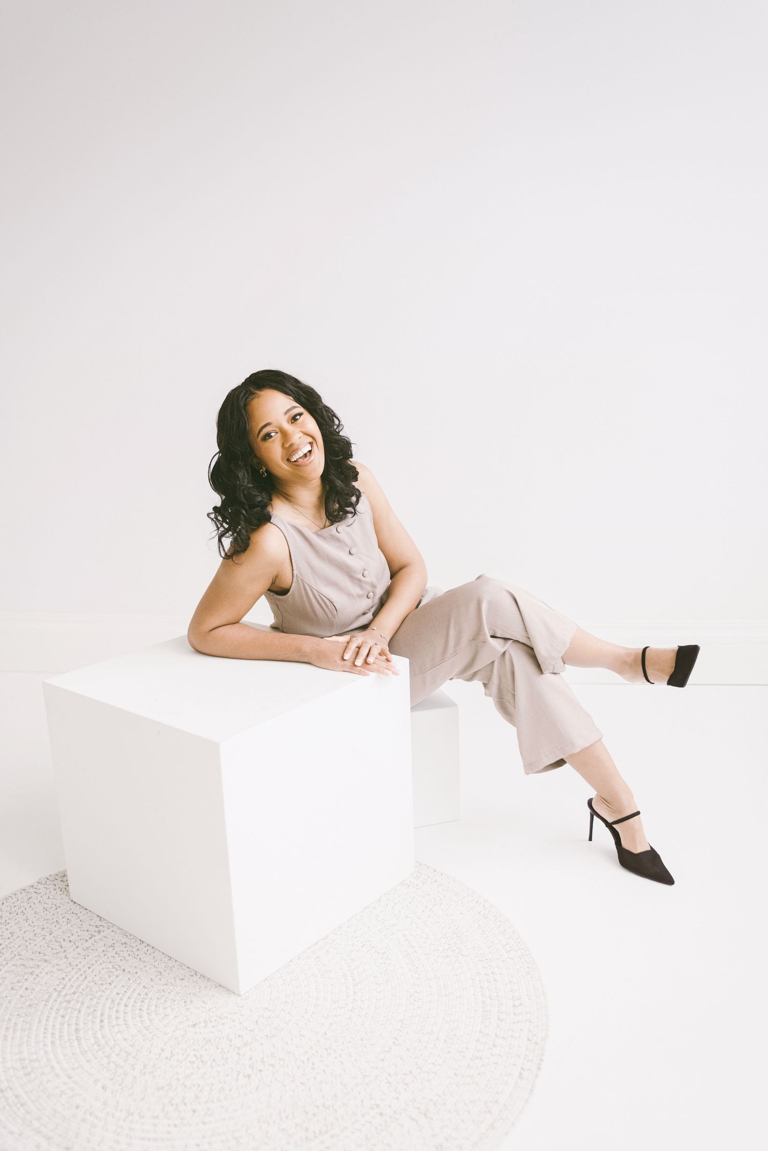 Smiling woman in beige jumpsuit and black heels sitting on white cube in bright studio setting.